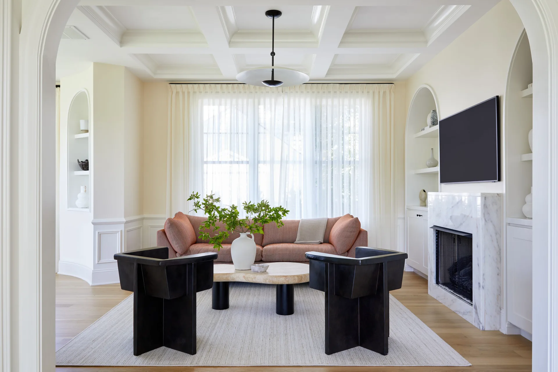 A bright living room features a coral sofa, black geometric armchairs, and an arched built-in fireplace.