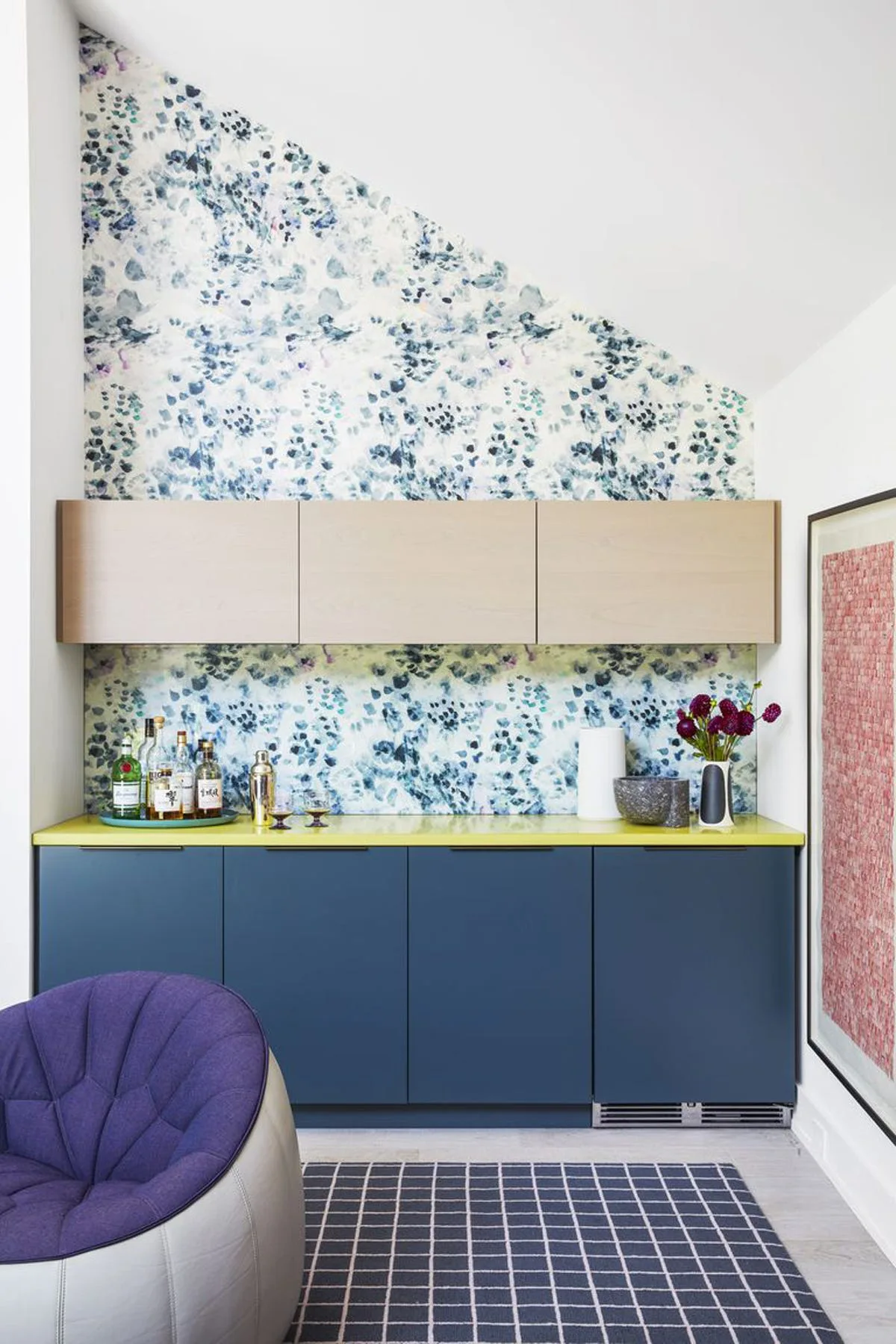 A home bar features deep blue lower cabinets with a bright yellow countertop, light wood upper cabinets, and floral wallpaper on a sloped wall.
