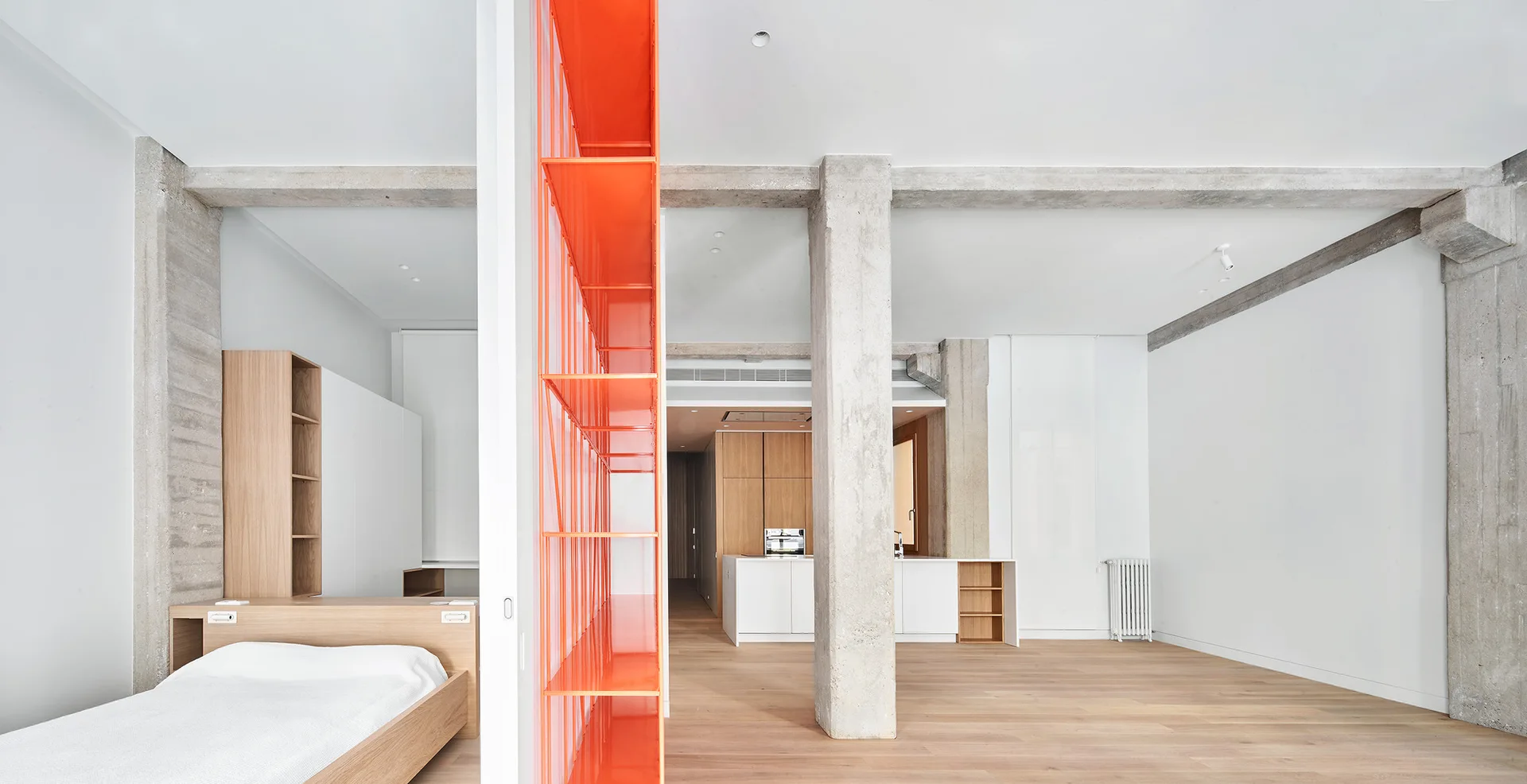 A minimalist open-plan apartment features exposed concrete columns, light wood floors, a low wooden bed, and a bright orange metal shelving unit.