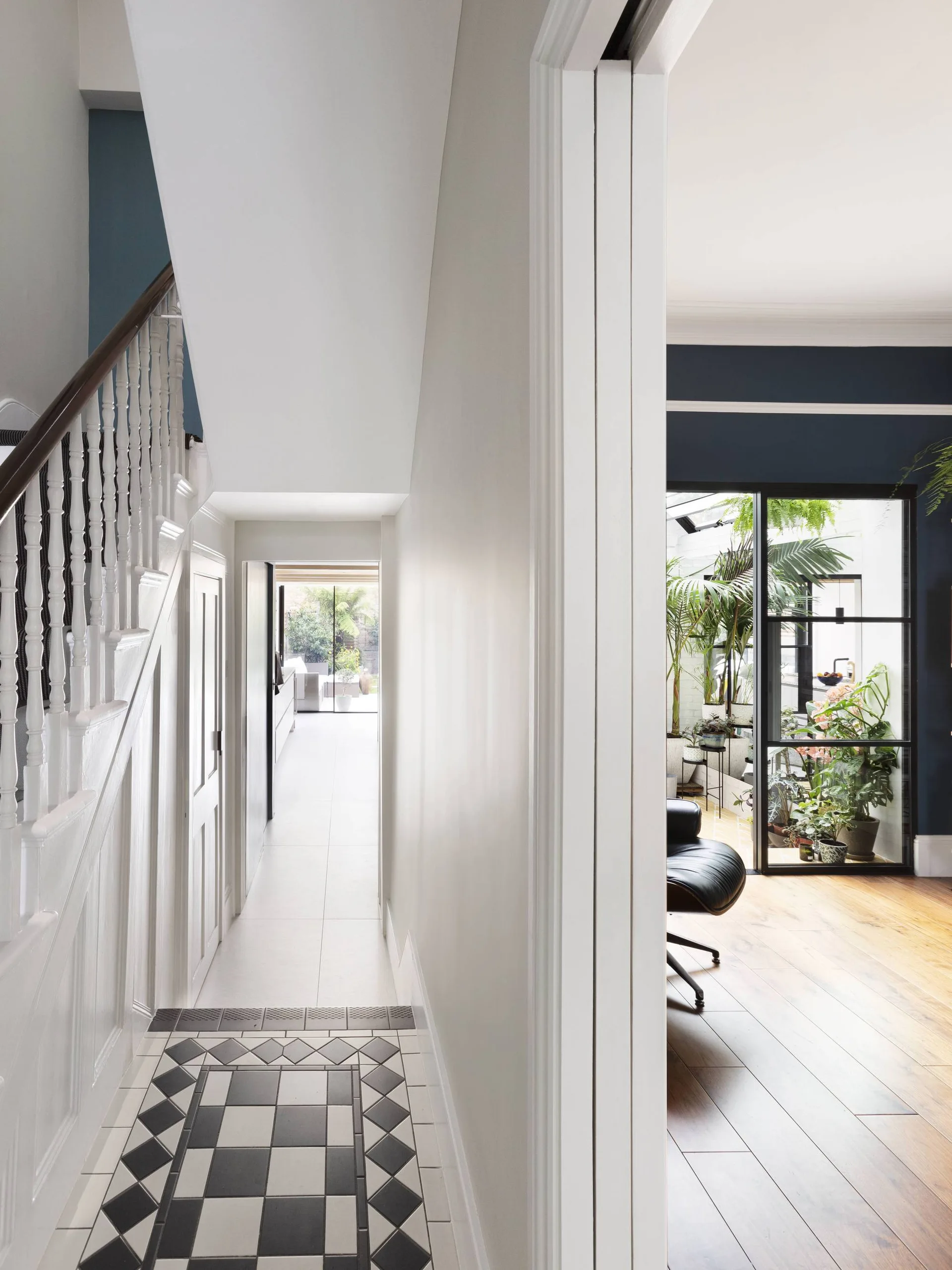 A bright white hallway with geometric black and white floor tiles leads past a staircase toward a sunroom framed by a black glass partition.
