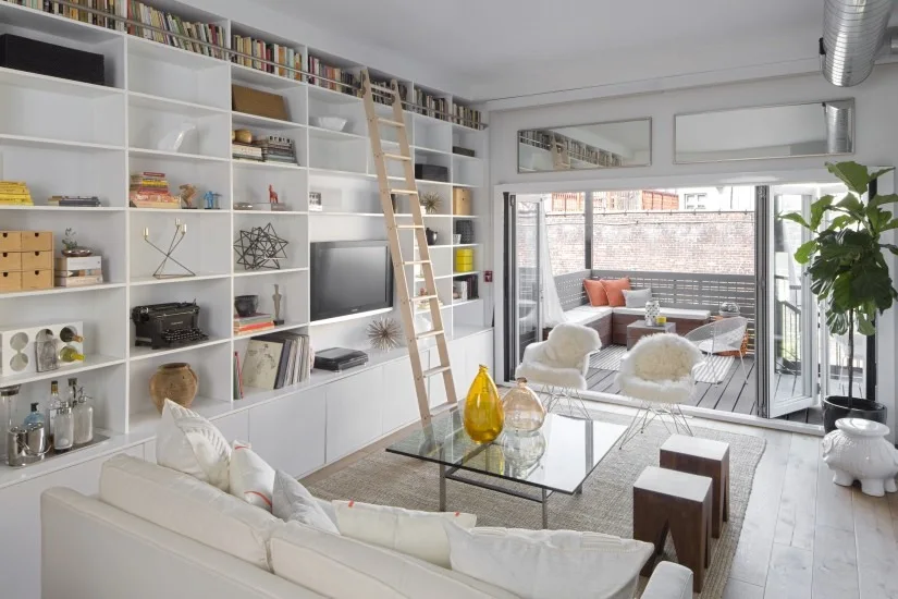 A bright living room with a white wall-to-wall bookcase and a sliding wooden ladder near bifold doors that open to a patio.