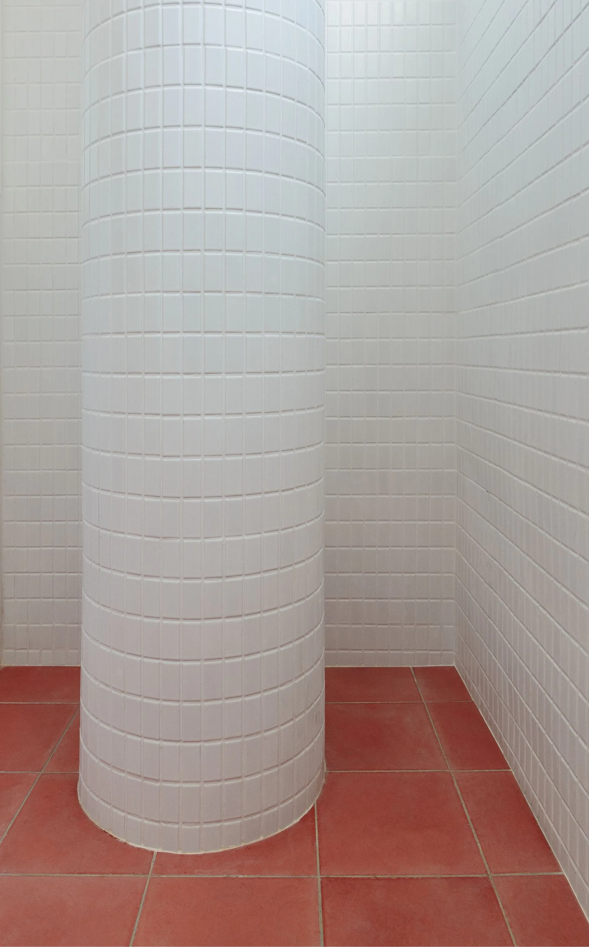 A rounded architectural column clad in white square tiles stands on a terracotta red tiled floor against a white tiled wall.