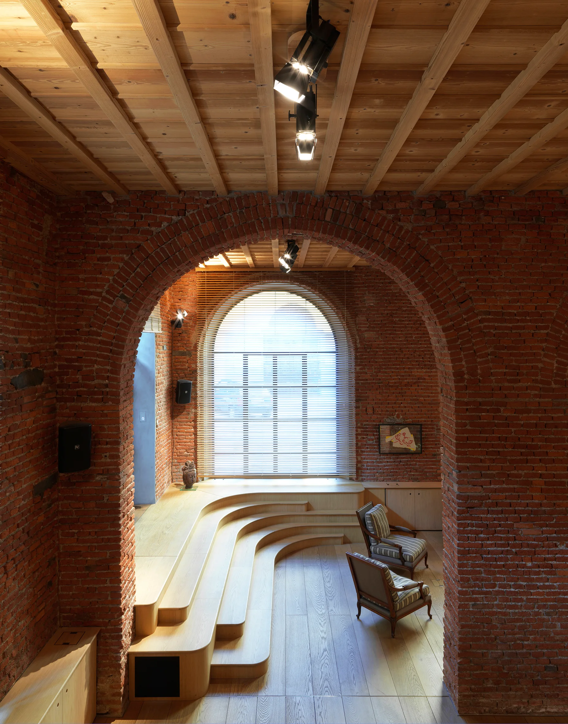 An interior view of a room with an exposed red brick archway, a multi-level stepped wood platform, and two armchairs under a wood ceiling.