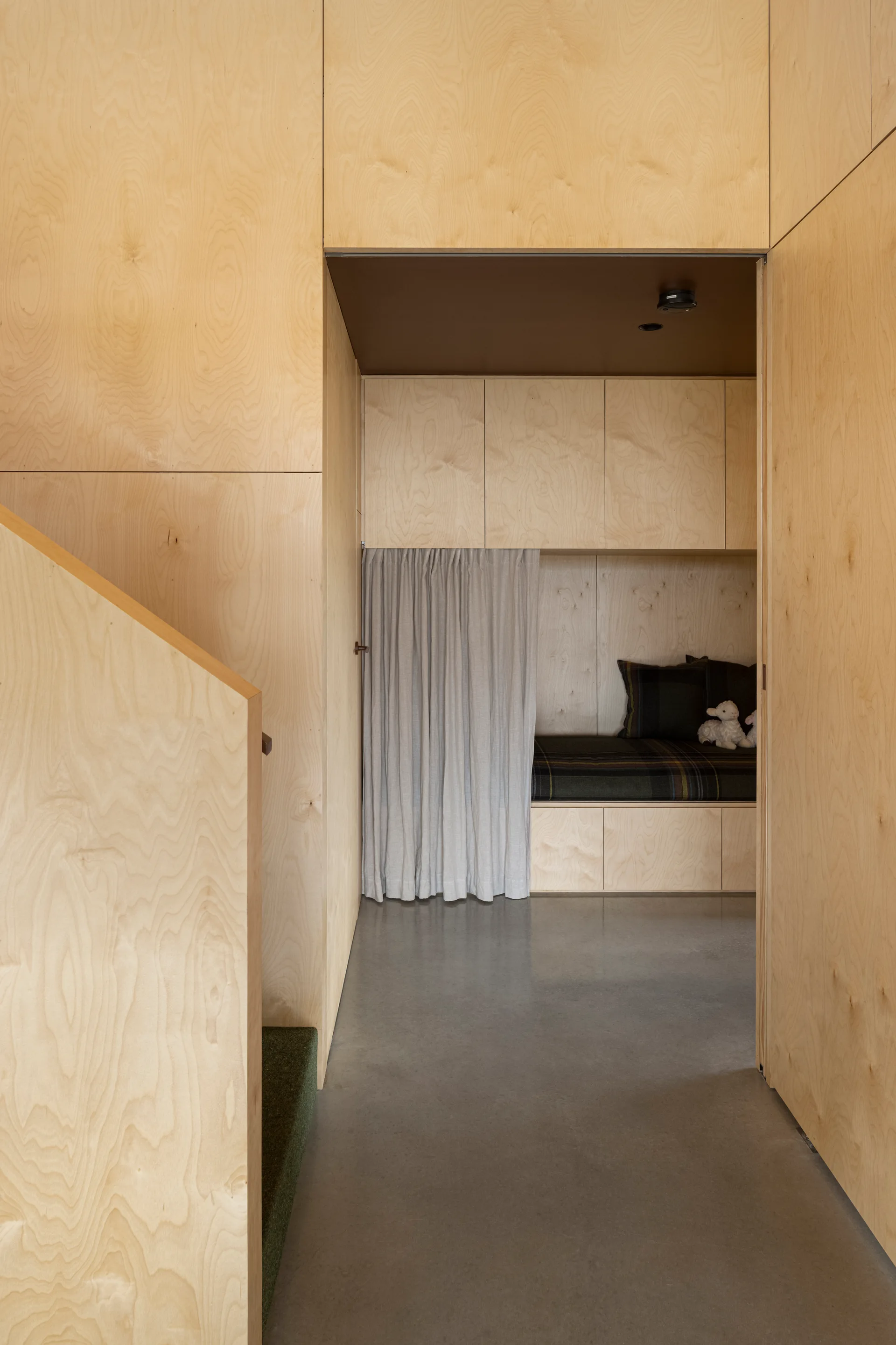 A built-in plywood sleeping nook with a grey curtain and plaid bedding is viewed through a hallway with polished concrete flooring.