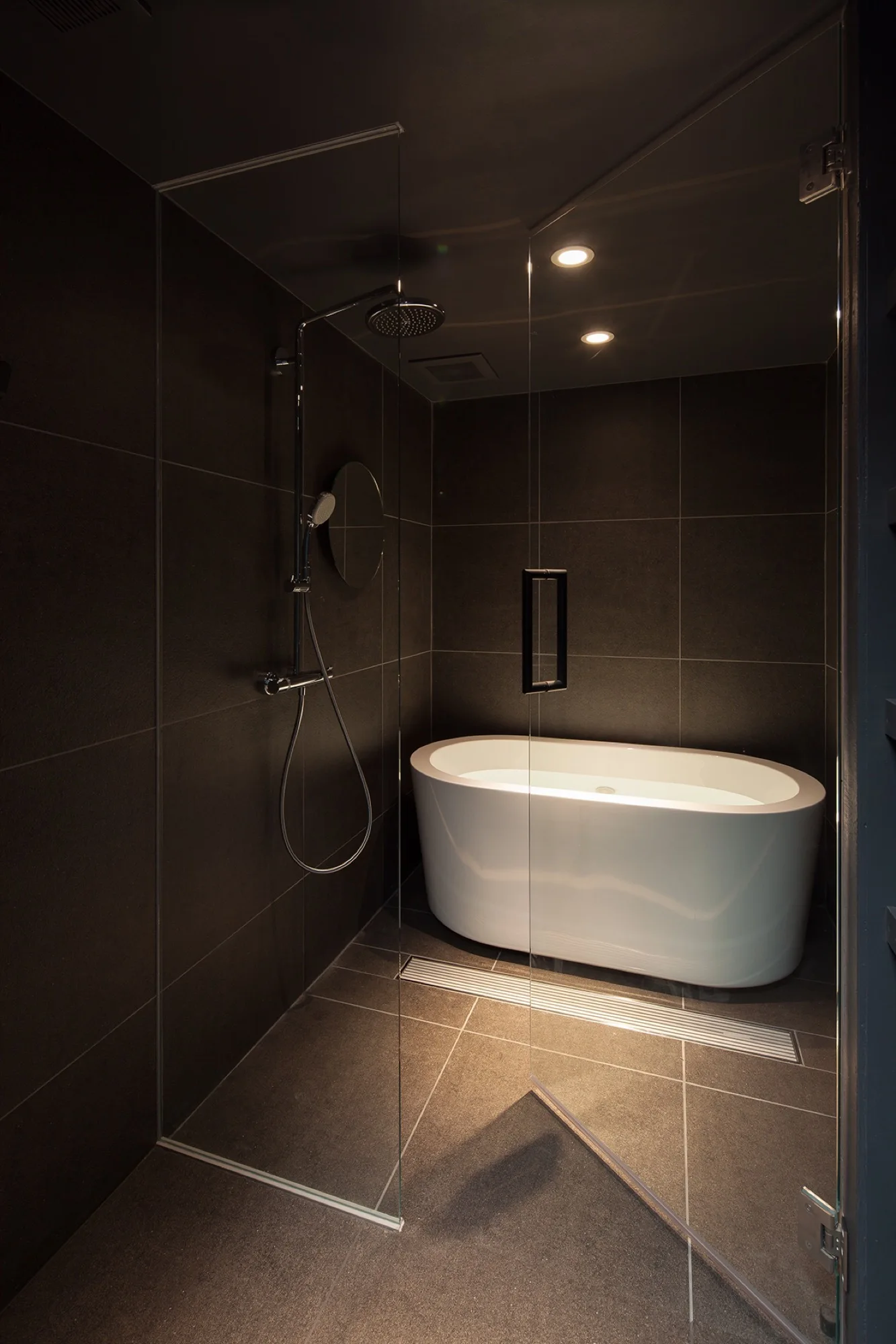 A modern wet room featuring dark gray tiled walls and floor, a white freestanding oval bathtub, and a glass shower enclosure.