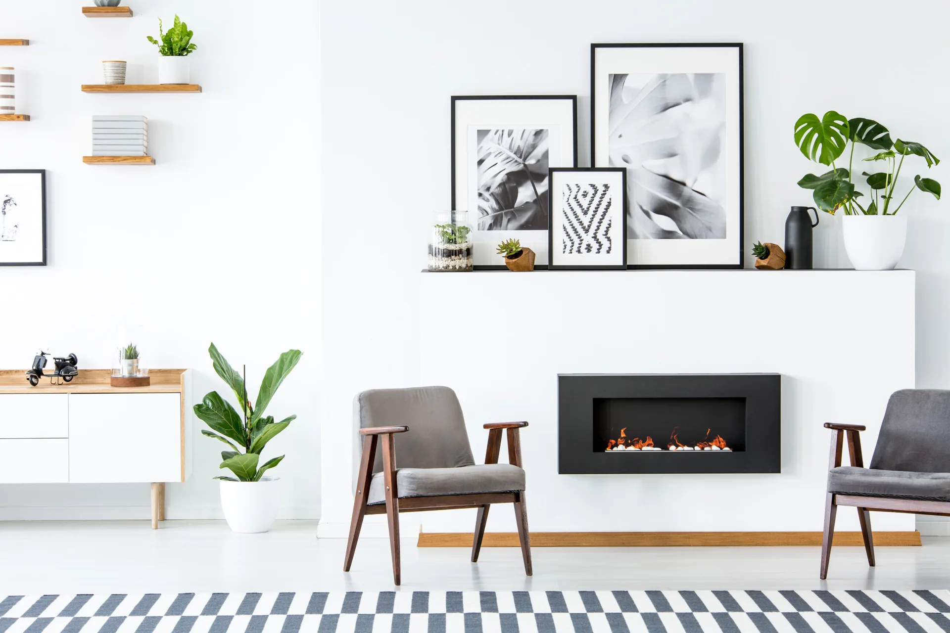 Two gray mid-century modern armchairs face a black inset fireplace on a white wall styled with layered framed botanical prints and green houseplants.