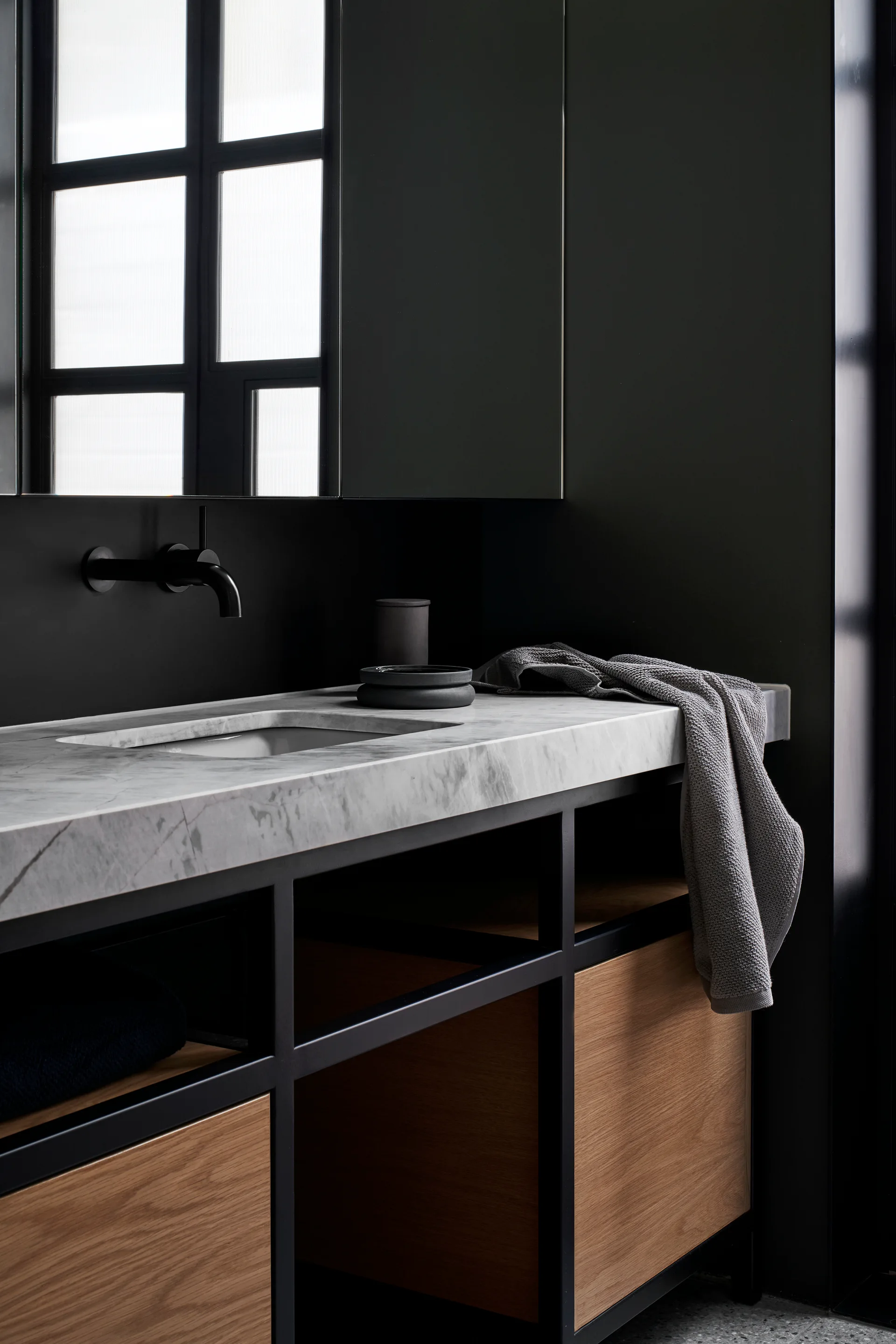 A dark bathroom vanity features a veined white marble countertop, a matte black wall-mounted faucet, and natural wood drawer fronts.