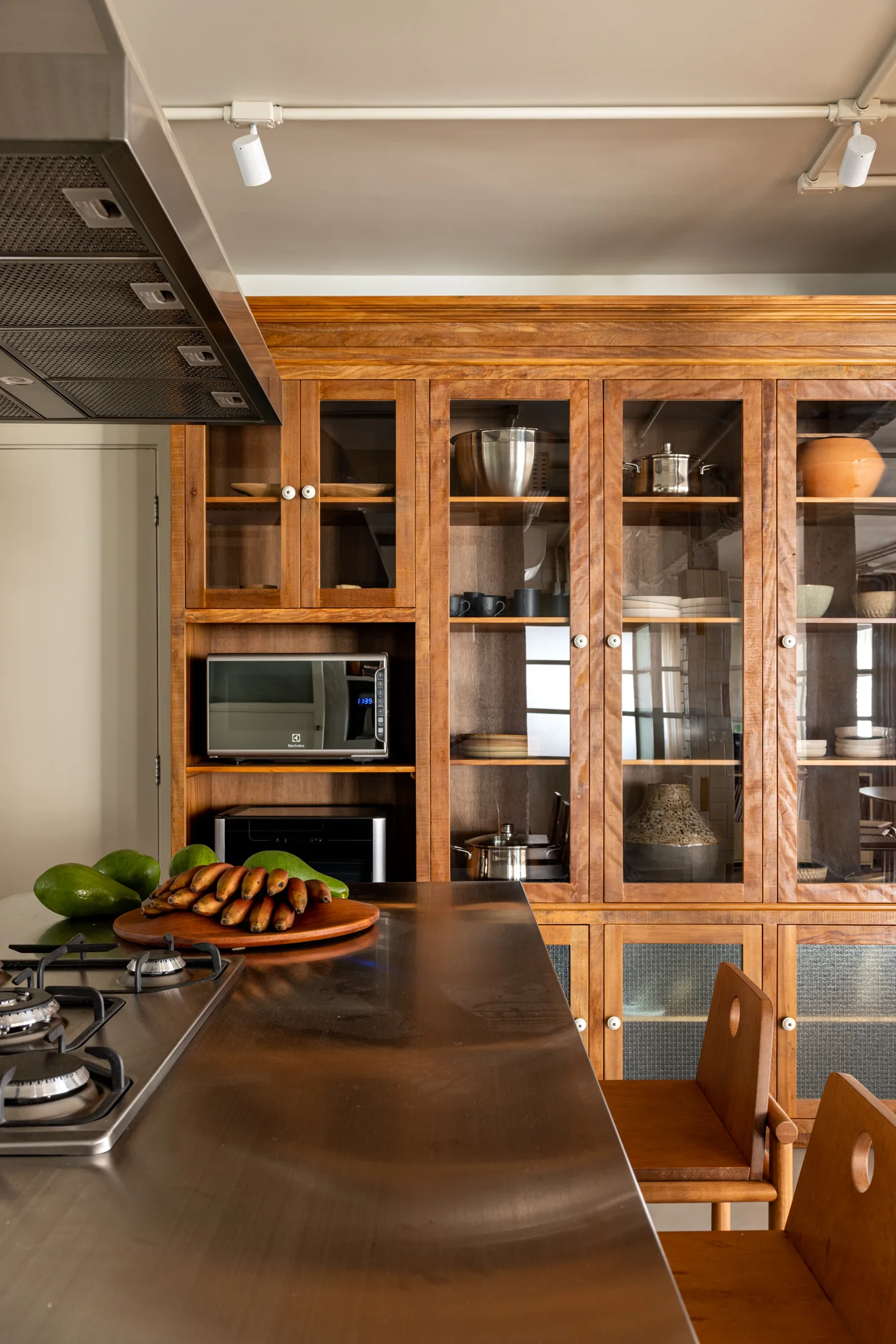 A kitchen features a stainless steel island with a gas cooktop opposite a large warm wood cabinet unit with glass doors.