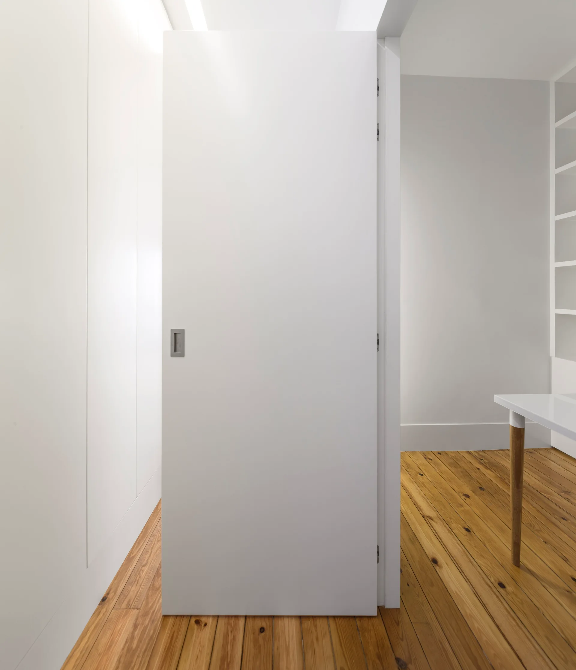 A minimalist white interior door with a recessed metal handle stands open on knotty pine wood floors.