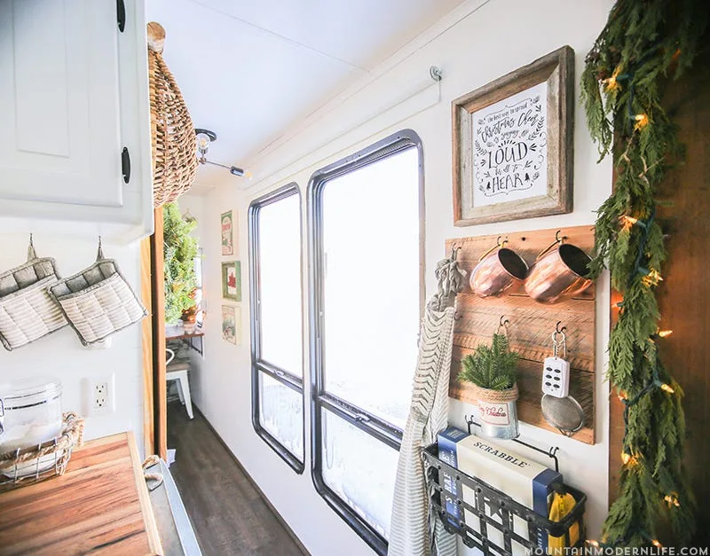A narrow white kitchen wall features a wooden rack hanging copper mugs alongside a Christmas garland and large vertical windows.