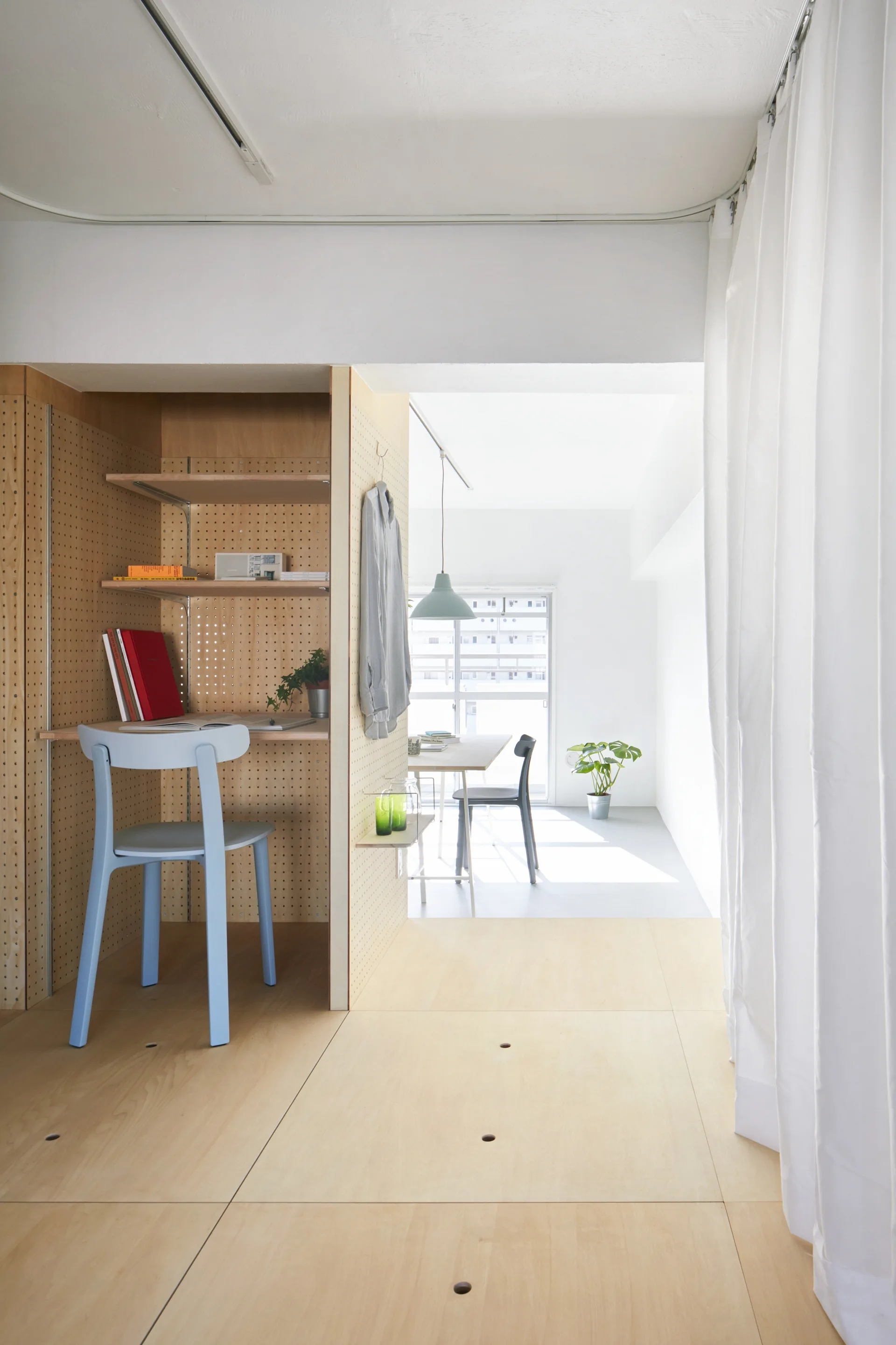 A minimalist plywood home office nook features a built-in desk with pegboard walls, a light blue chair, and a white curtain partition.
