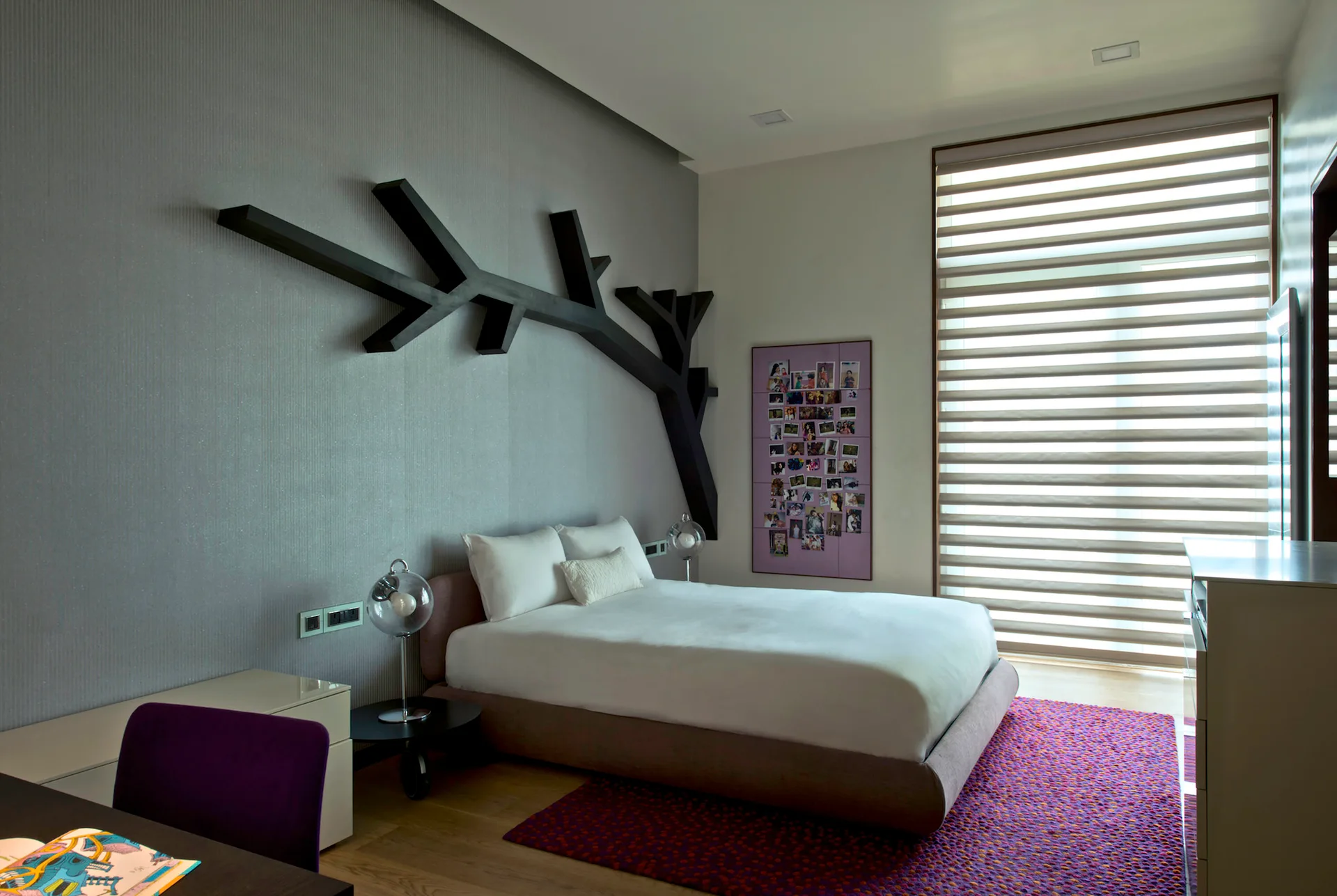 A contemporary bedroom features a bed with white linens, a black tree branch wall sculpture on a grey textured wall, and a purple area rug.