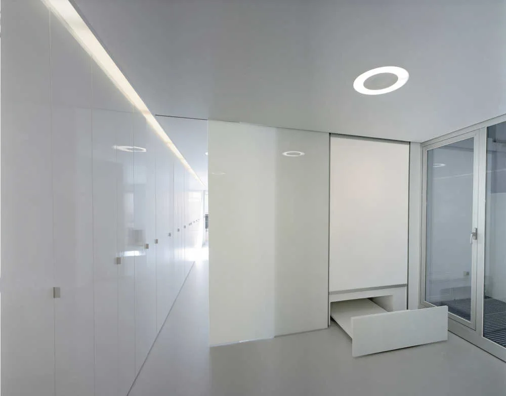 A minimalist white hallway featuring seamless floor-to-ceiling cabinets, a recessed circular ceiling light, a linear cove light, and a glass partition door.