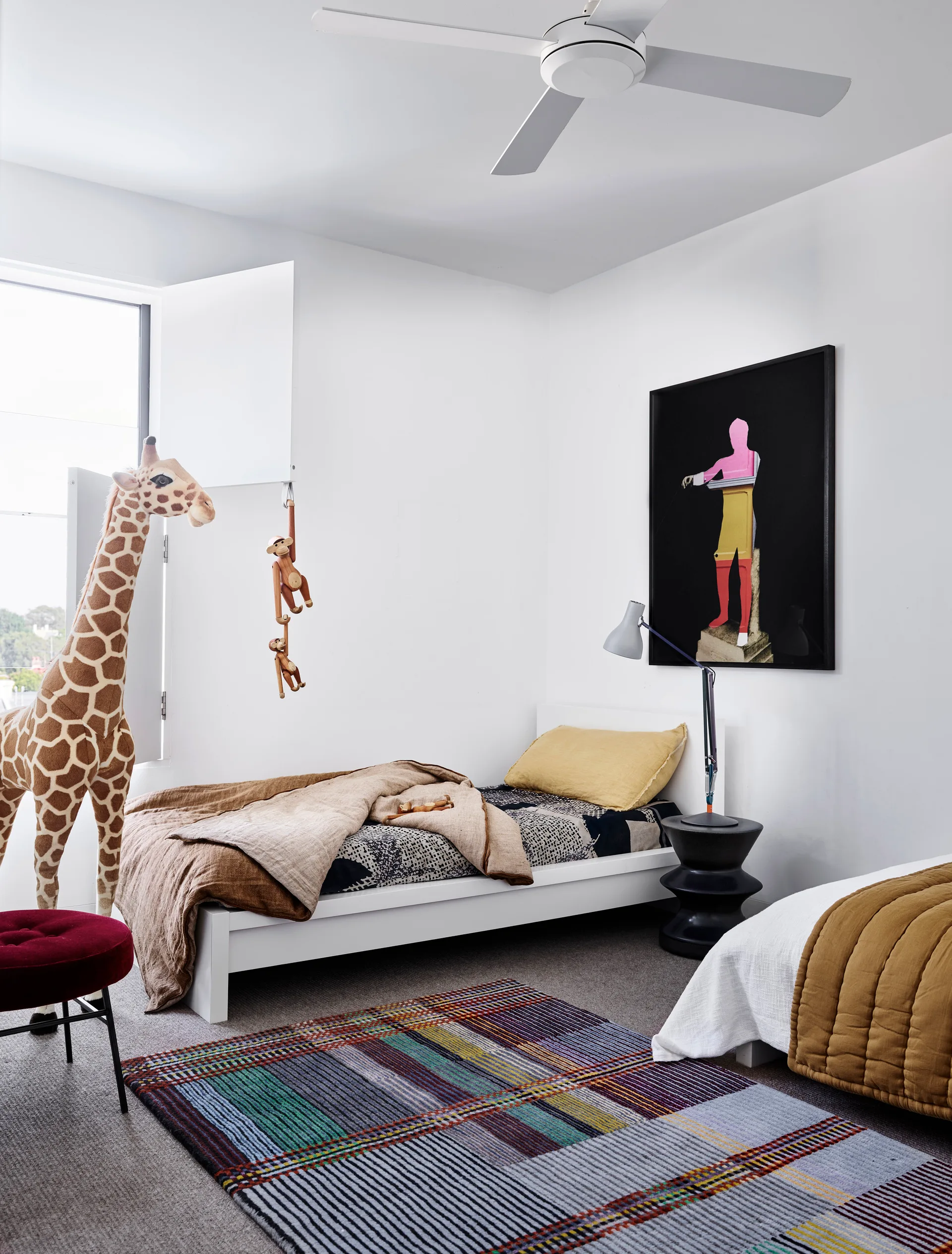 A modern kids bedroom features a white twin bed, a large stuffed giraffe by a shuttered window, a colorful plaid area rug, and a graphic framed art print.