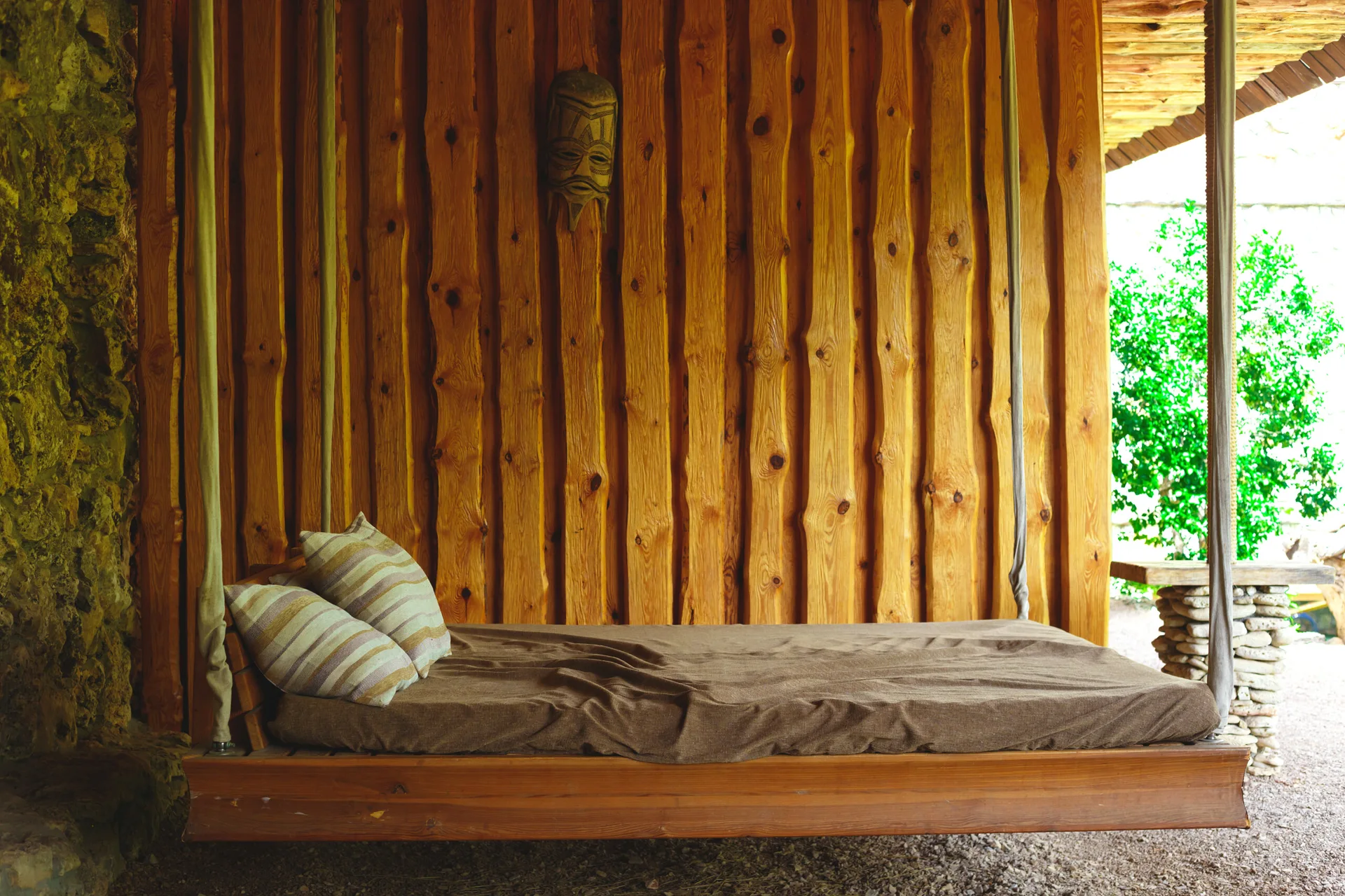A wooden hanging daybed with striped pillows is suspended in front of a vertical wood plank wall on a rustic covered patio.