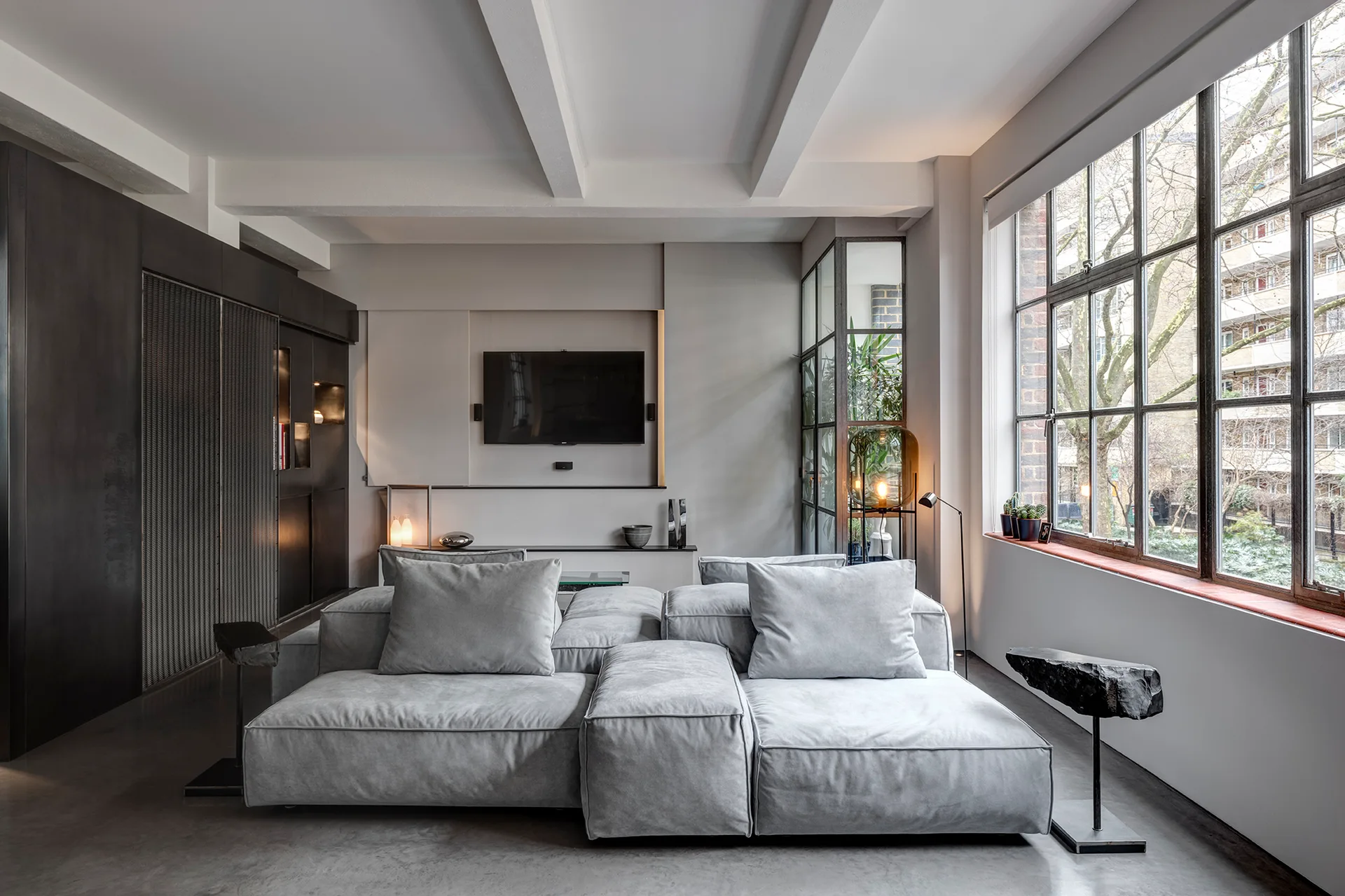 A minimalist living room featuring a grey modular sectional sofa, concrete flooring, and a large black grid industrial window showing outdoor trees.