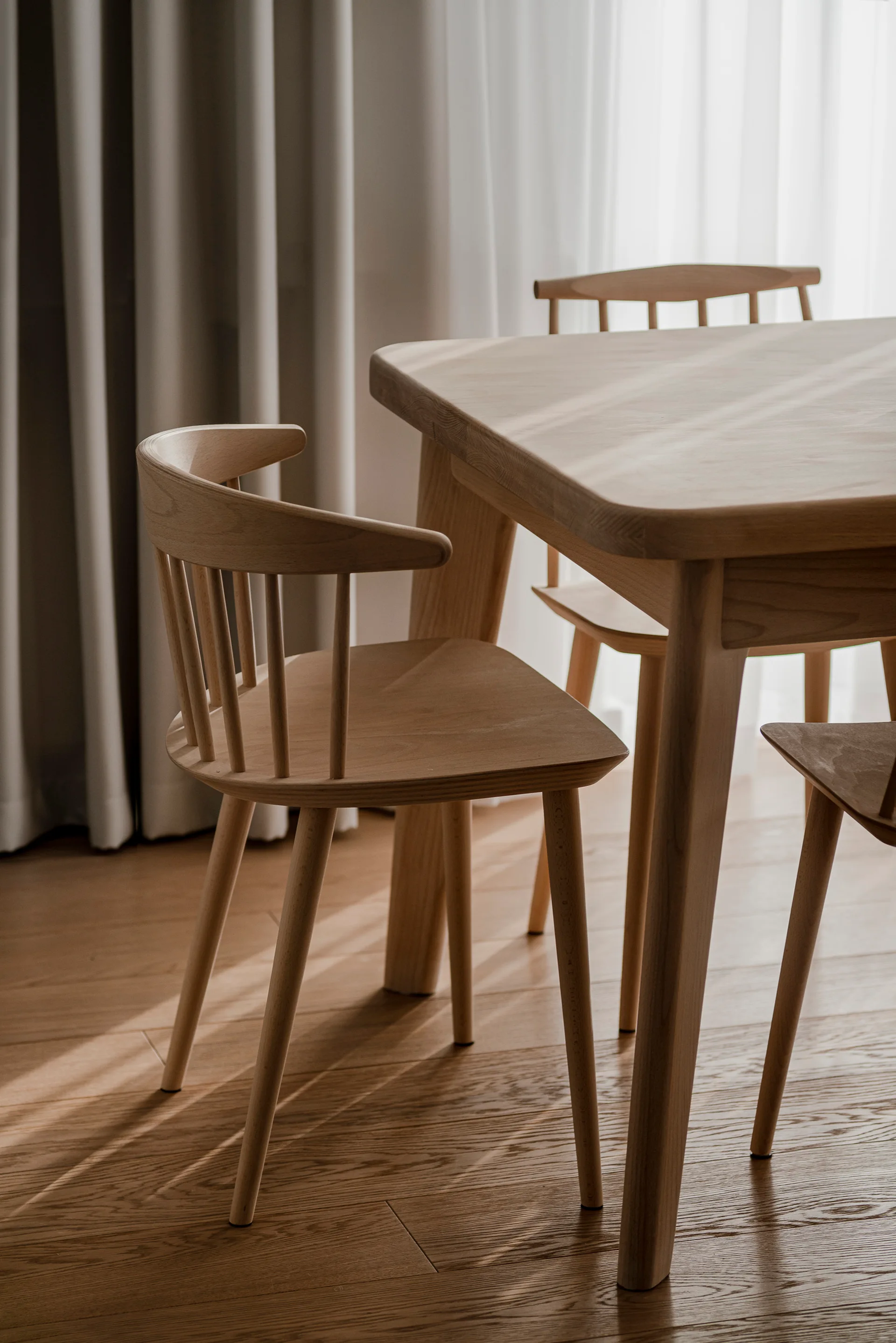 A light wood dining table and matching spindle-back wooden chairs sit on a wood plank floor beside sheer white curtains in soft daylight.
