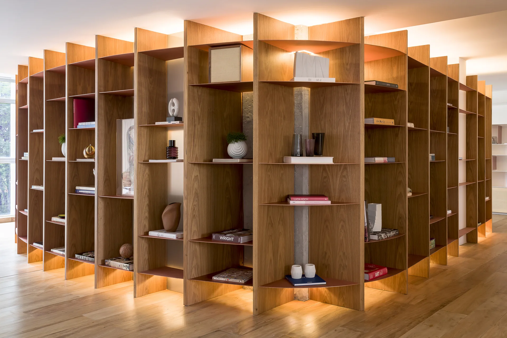 A custom wooden shelving unit with tall vertical fins and integrated warm lighting stands on light hardwood floors, displaying books and decorative objects.