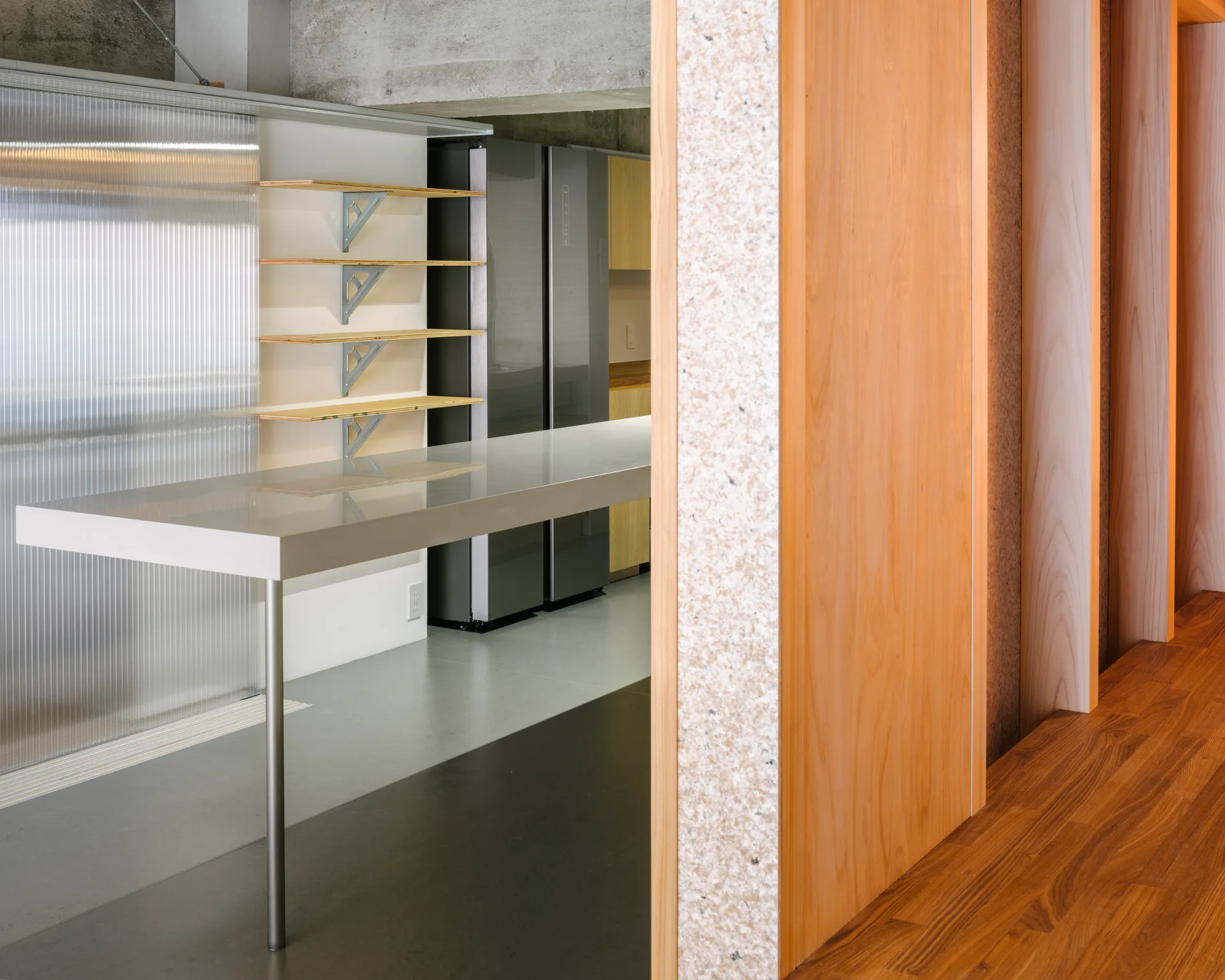 A modern industrial kitchen features a grey island countertop beneath an exposed concrete ceiling, with ribbed wall panels and warm wood partitions nearby.