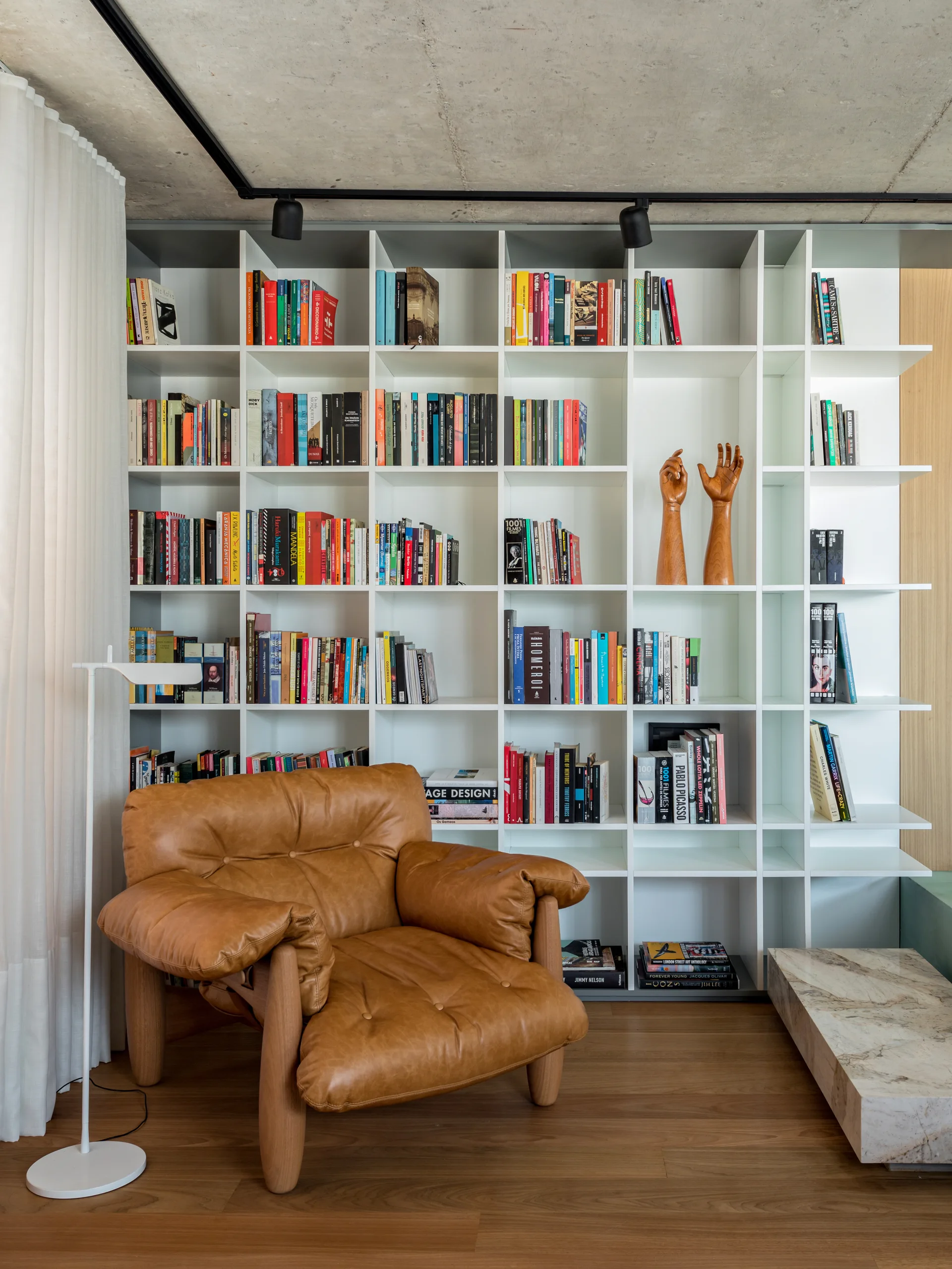 A brown leather armchair sits on wood flooring in front of a white grid bookshelf filled with books beneath an exposed concrete ceiling.
