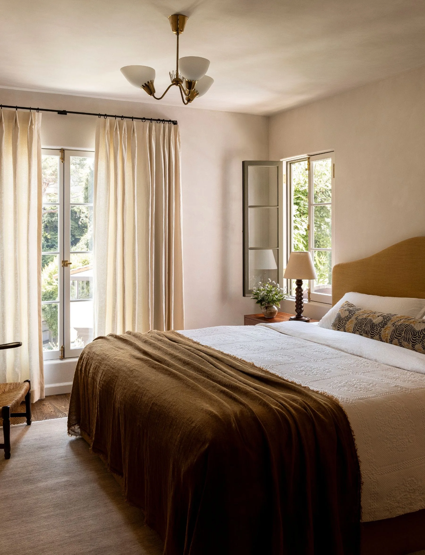 A bedroom features a bed with an ochre headboard and a brown throw, flanked by casement windows with long cream curtains.