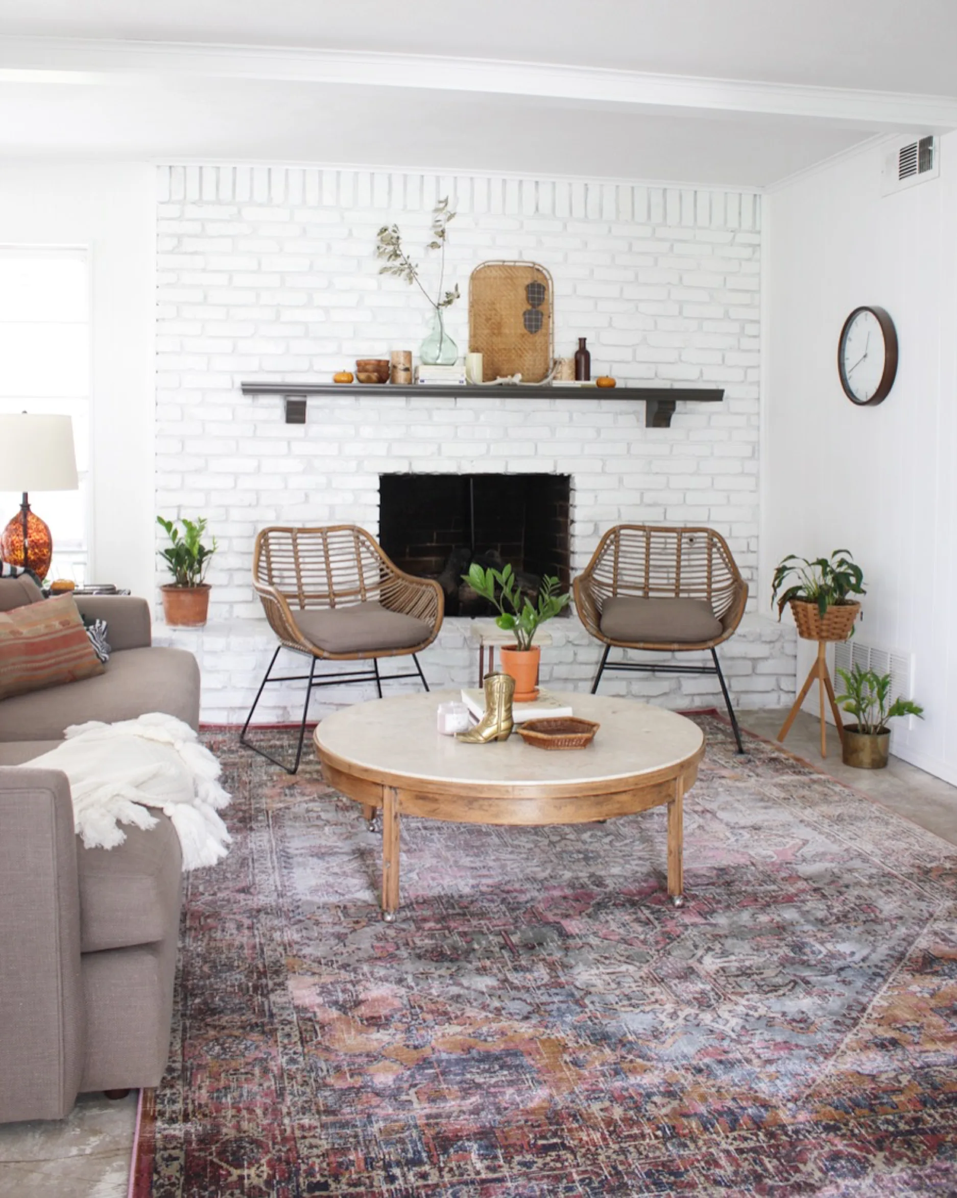 A living room features a white painted brick fireplace with a dark wood mantel, two rattan armchairs, and a large patterned area rug.