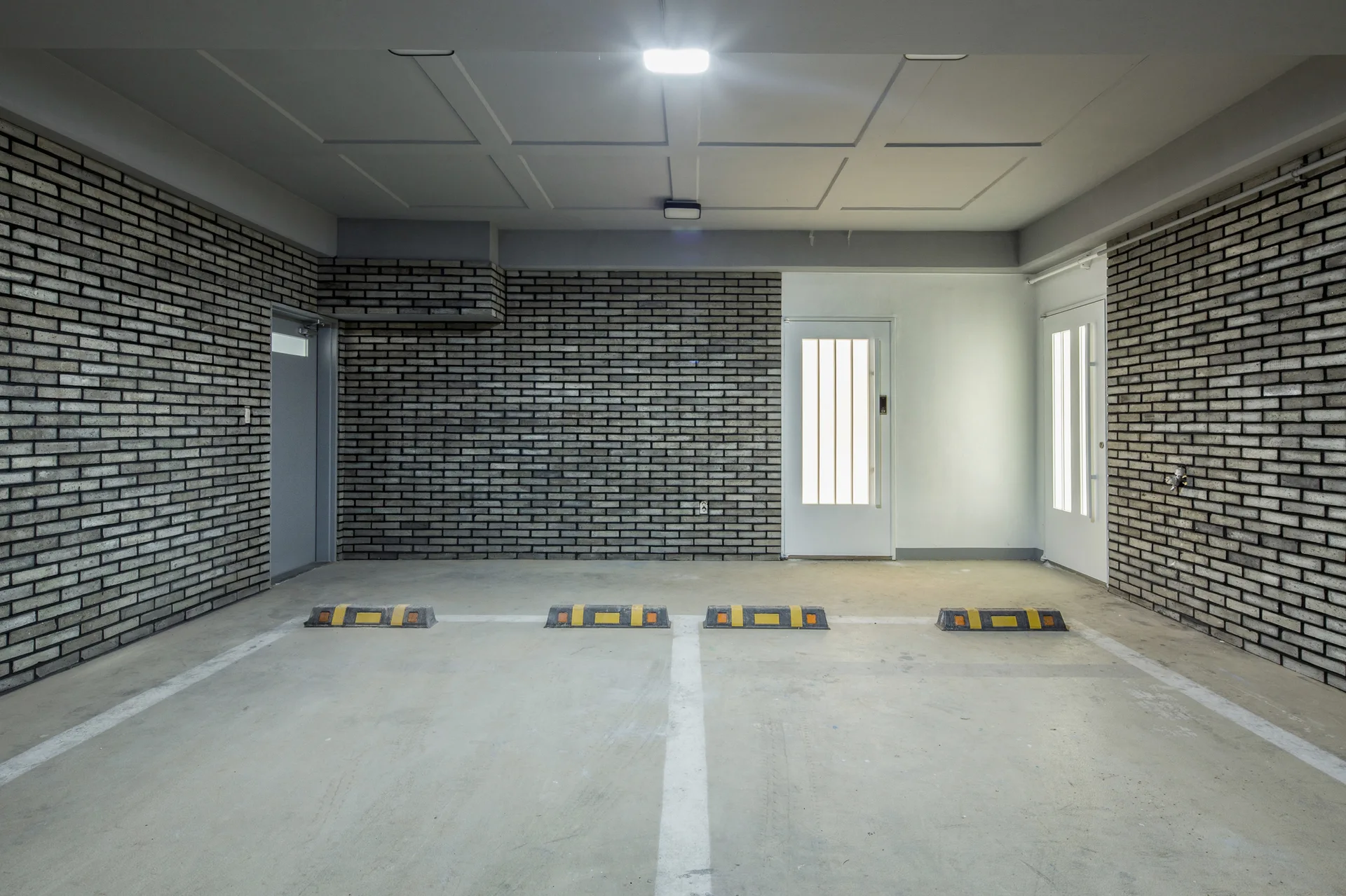 An indoor parking garage features dark gray brick walls, a paneled gray ceiling, white doors, and a concrete floor with rubber wheel stops.