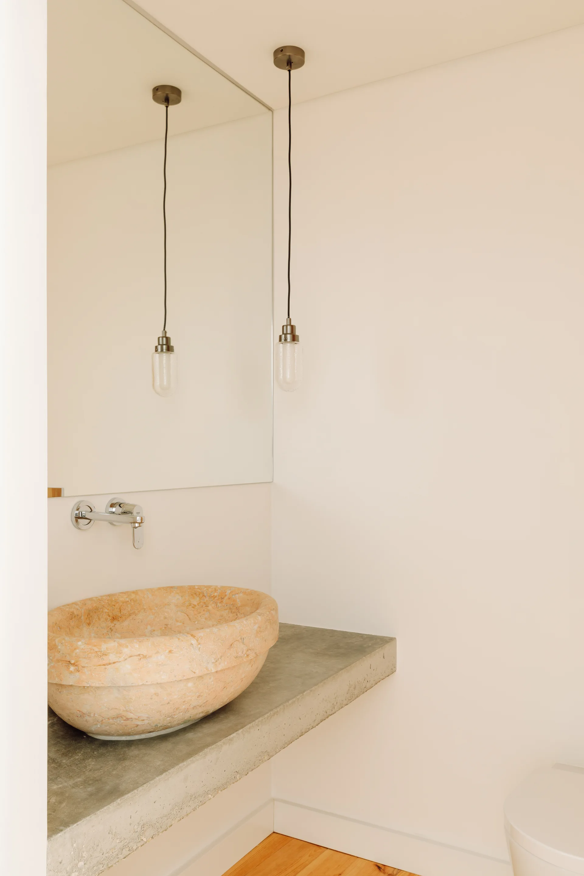 A minimalist bathroom features a textured peach stone vessel sink on a floating raw concrete counter under a wall-mounted chrome faucet.