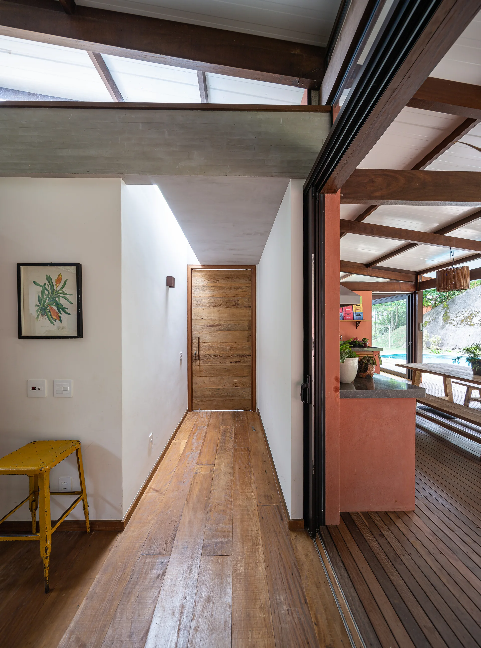 A bright hallway with light wood floors and a skylight leads past a terracotta-colored wall toward a rustic wood door.