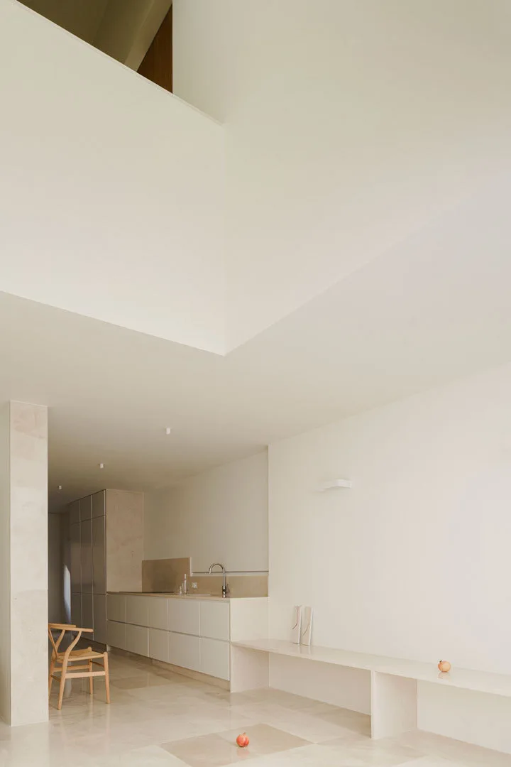 A minimalist off-white kitchen features flat-front cabinets, a low stone bench along the wall, and a light wood wishbone chair on stone tiled flooring.