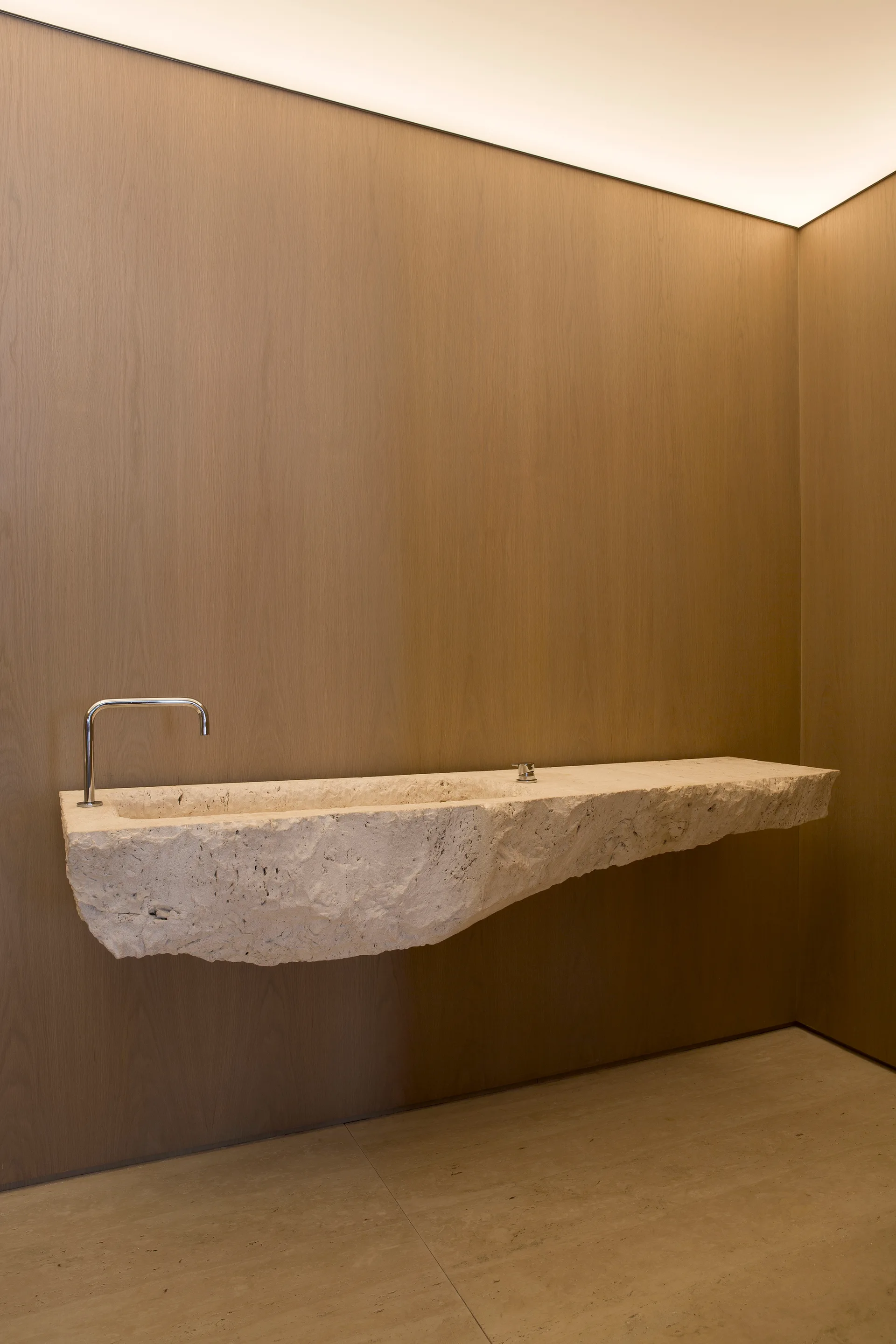 A wall-mounted rough-cut stone sink with a chrome faucet stands against vertical wood-paneled walls in a minimalist bathroom under soft cove lighting.