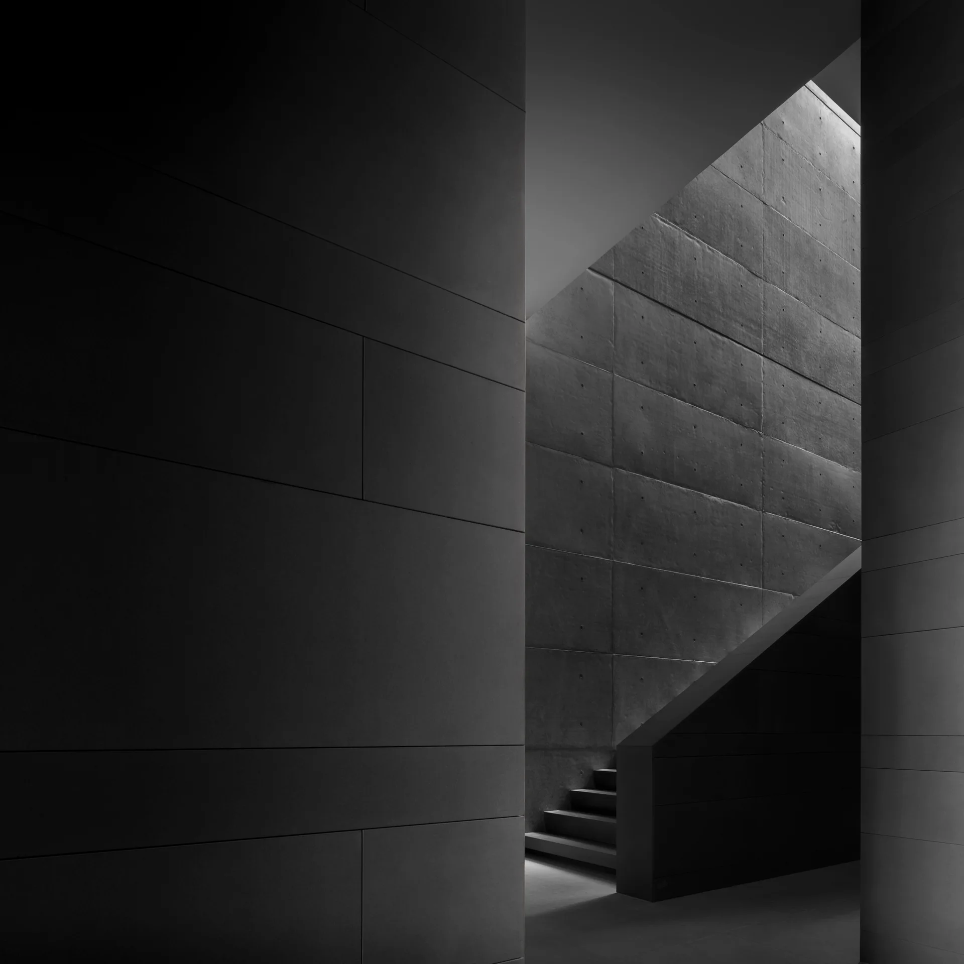 A minimalist concrete staircase rises alongside a gridded wall illuminated by a single shaft of light casting deep shadows.