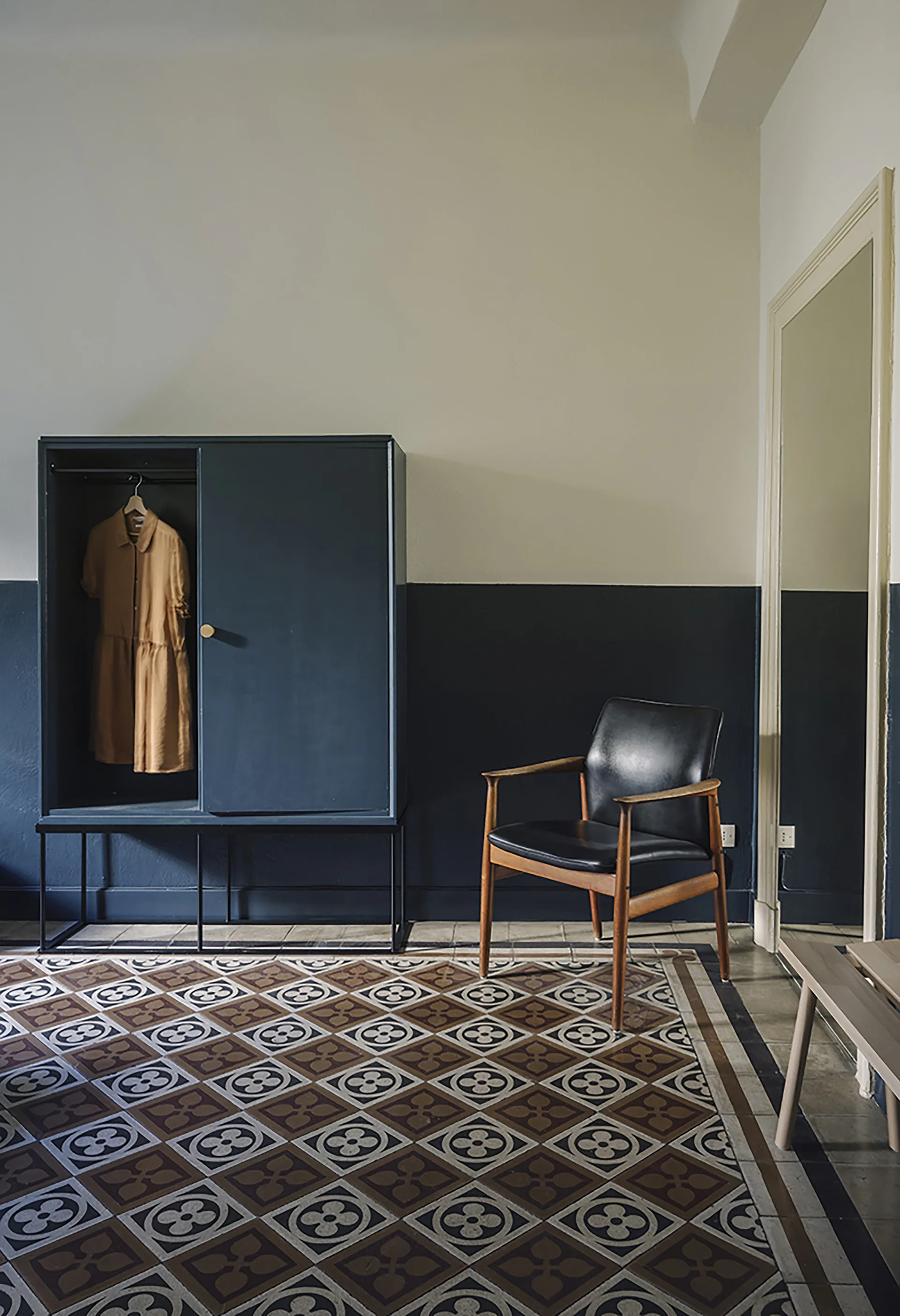 A dark wardrobe and a black leather armchair sit on geometric patterned floor tiles against a two-tone painted wall.