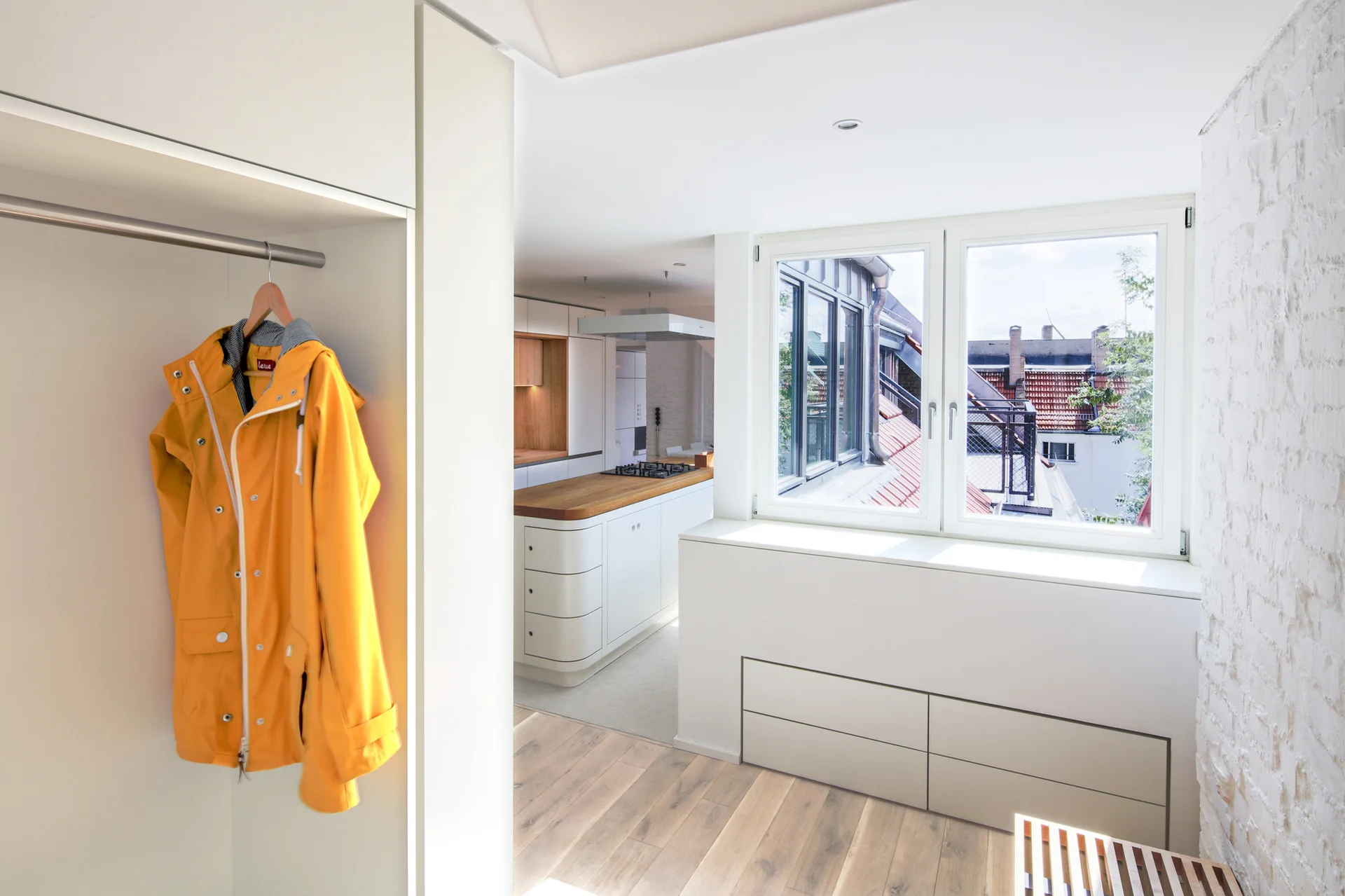 A bright entryway with white built-in cabinets and a yellow raincoat leads into a minimalist kitchen with a wood-topped island.