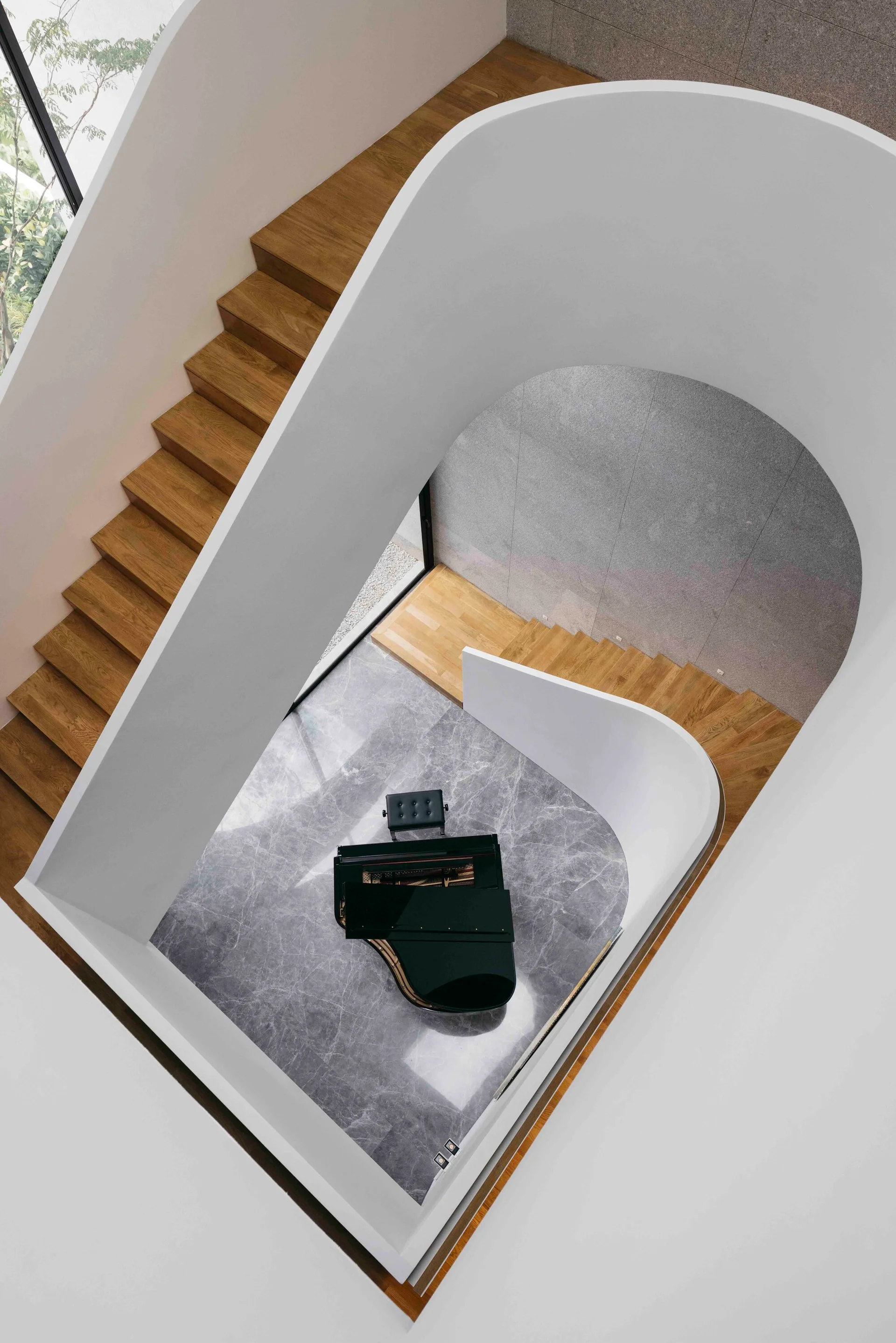 Looking down at a curved wooden staircase with white walls surrounding a black grand piano on a gray marbled floor.