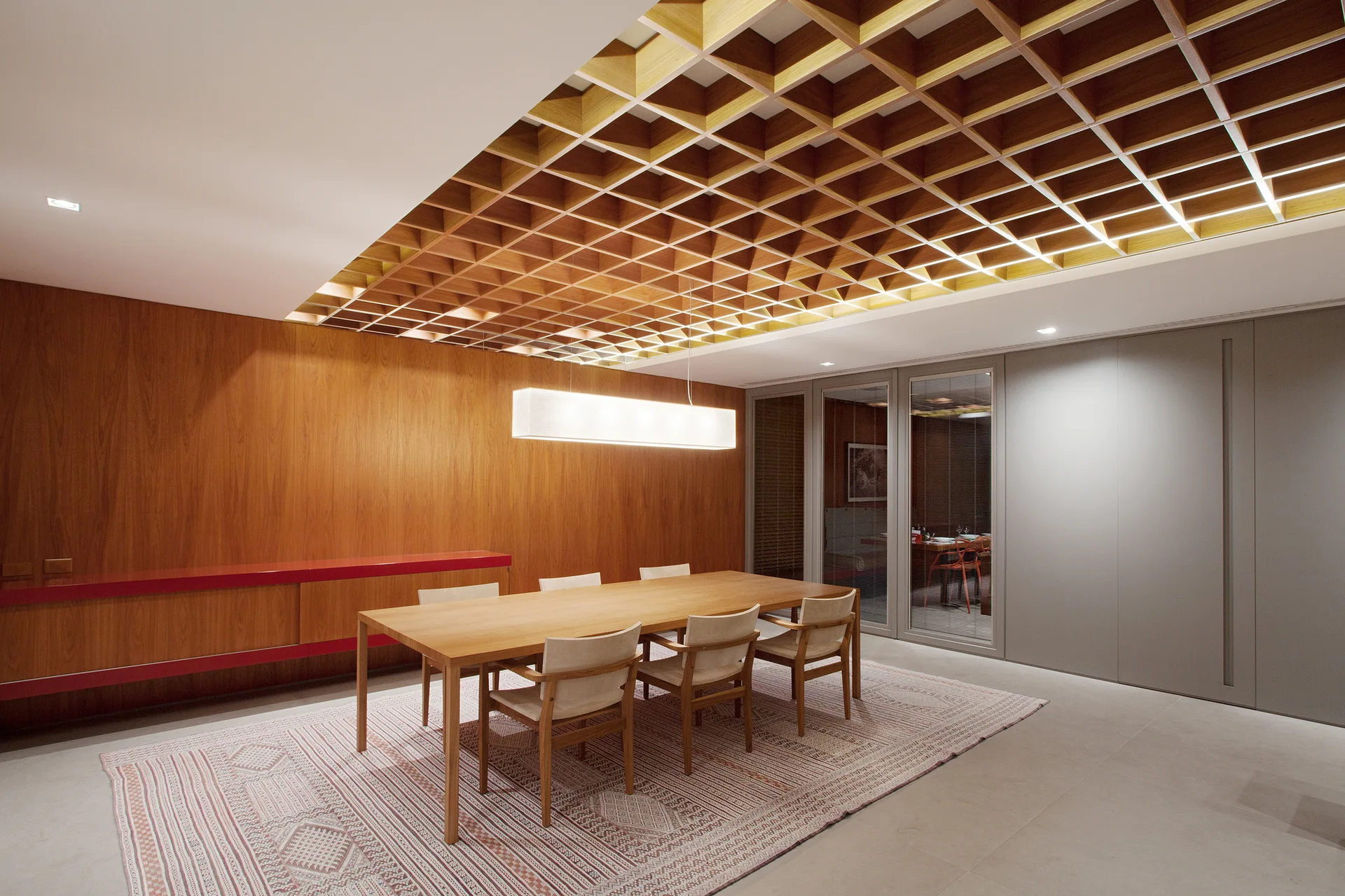 A modern dining room features a wooden table and chairs on a patterned rug below an intricate coffered wood ceiling and a red wall cabinet.