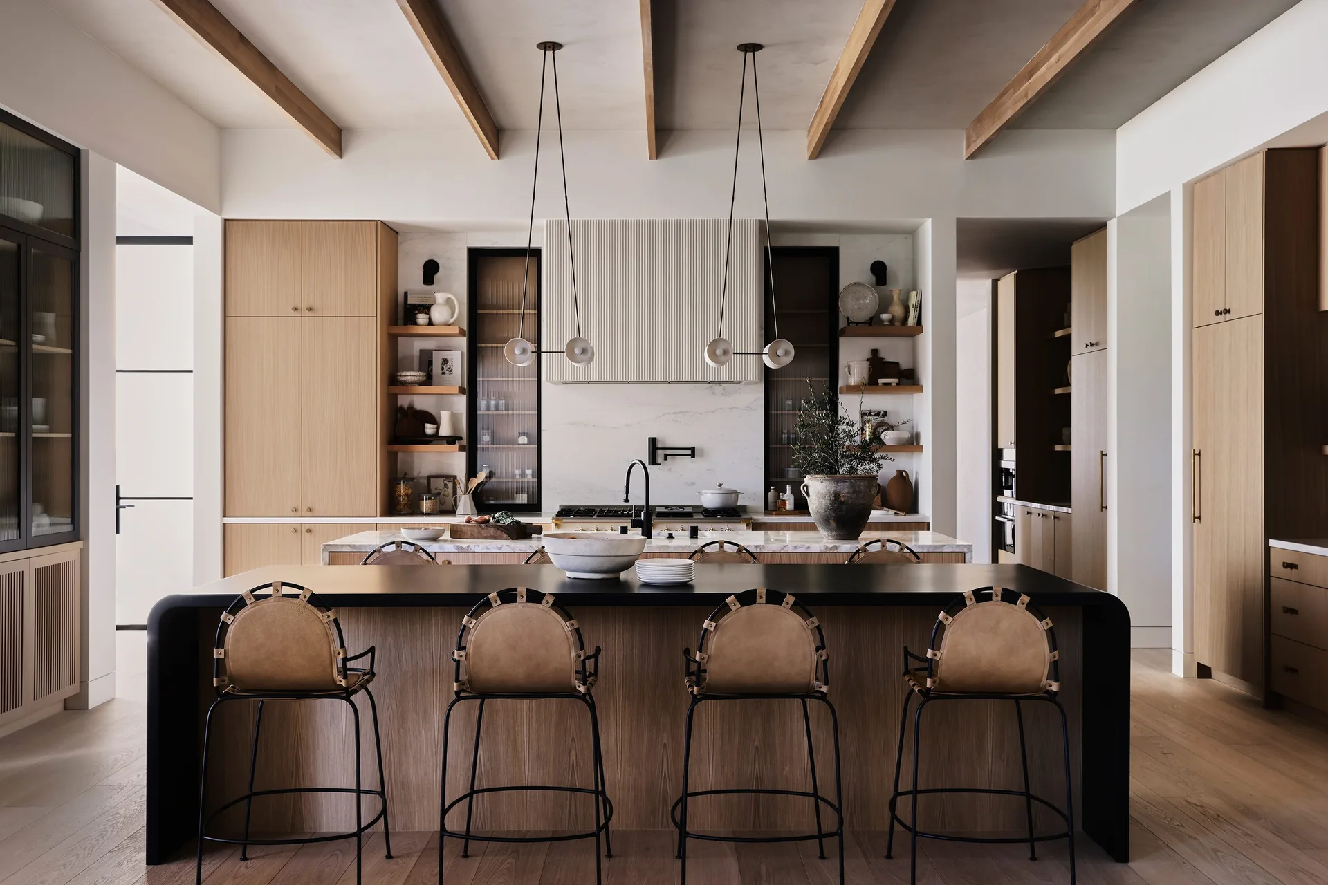 A modern kitchen with light oak cabinetry, exposed ceiling beams, a black-topped island with leather barstools, and a fluted range hood.