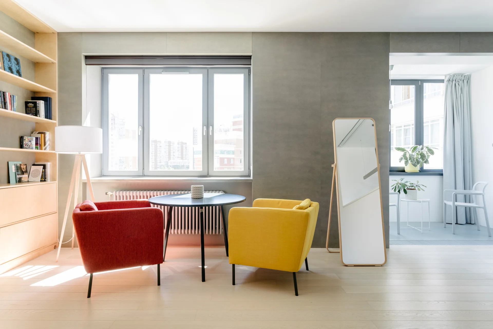 A minimalist living room with a round black table flanked by a yellow armchair and a red armchair in front of a large window.