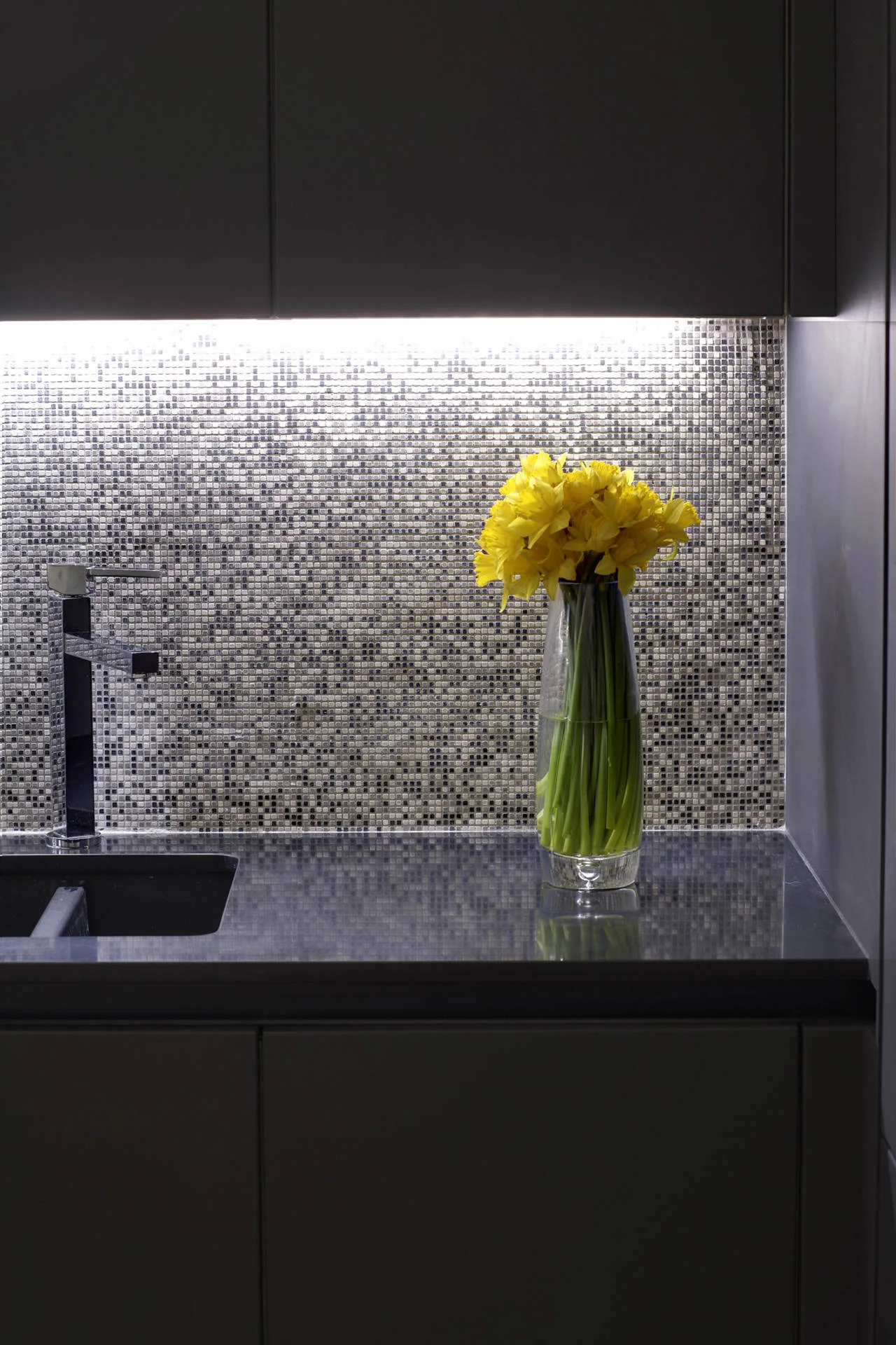 A dark gray kitchen counter features an integrated sink and a glass vase of yellow daffodils set against an illuminated mosaic tile backsplash.