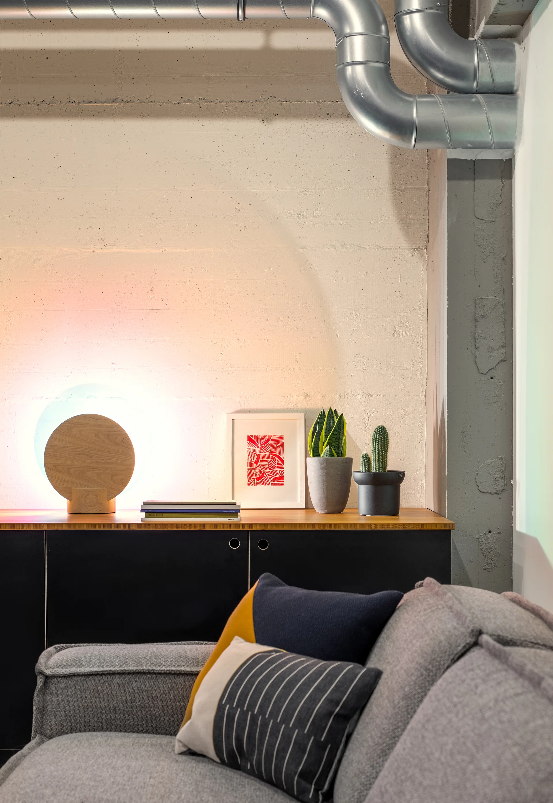 A grey sofa sits in front of a black credenza decorated with potted plants, framed art, and a circular wooden lamp against a textured concrete wall.