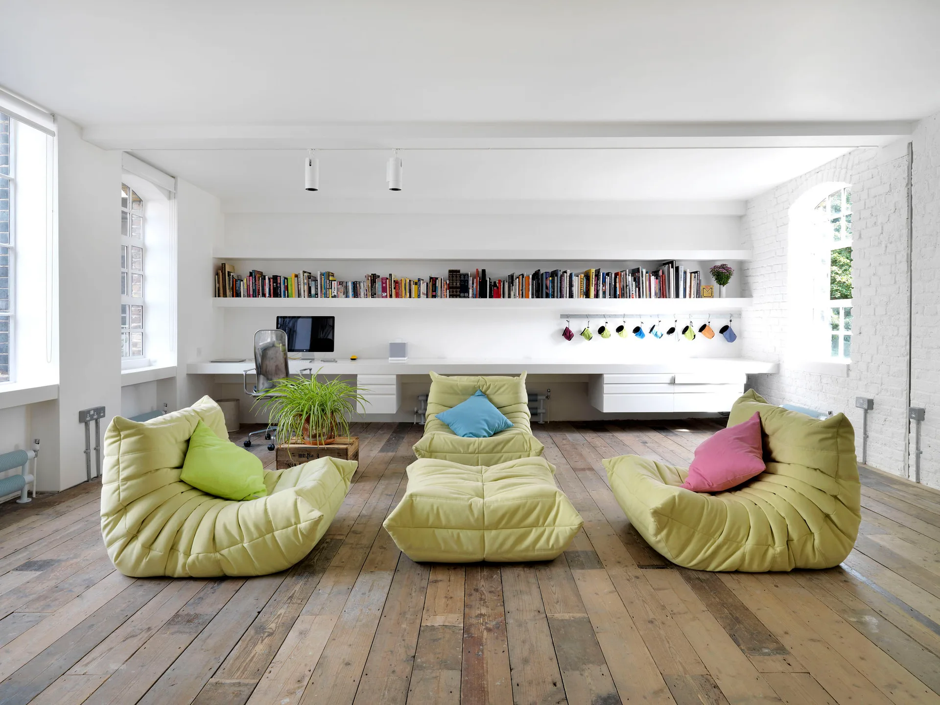 A bright white room features a long floating desk and matching bookshelf above three light green slouchy lounge chairs on a rustic wood floor.