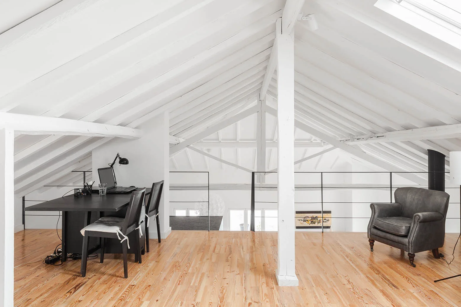 An attic office loft features a sloped white beamed ceiling, natural wood flooring, a black desk, and a grey armchair next to a black metal railing.