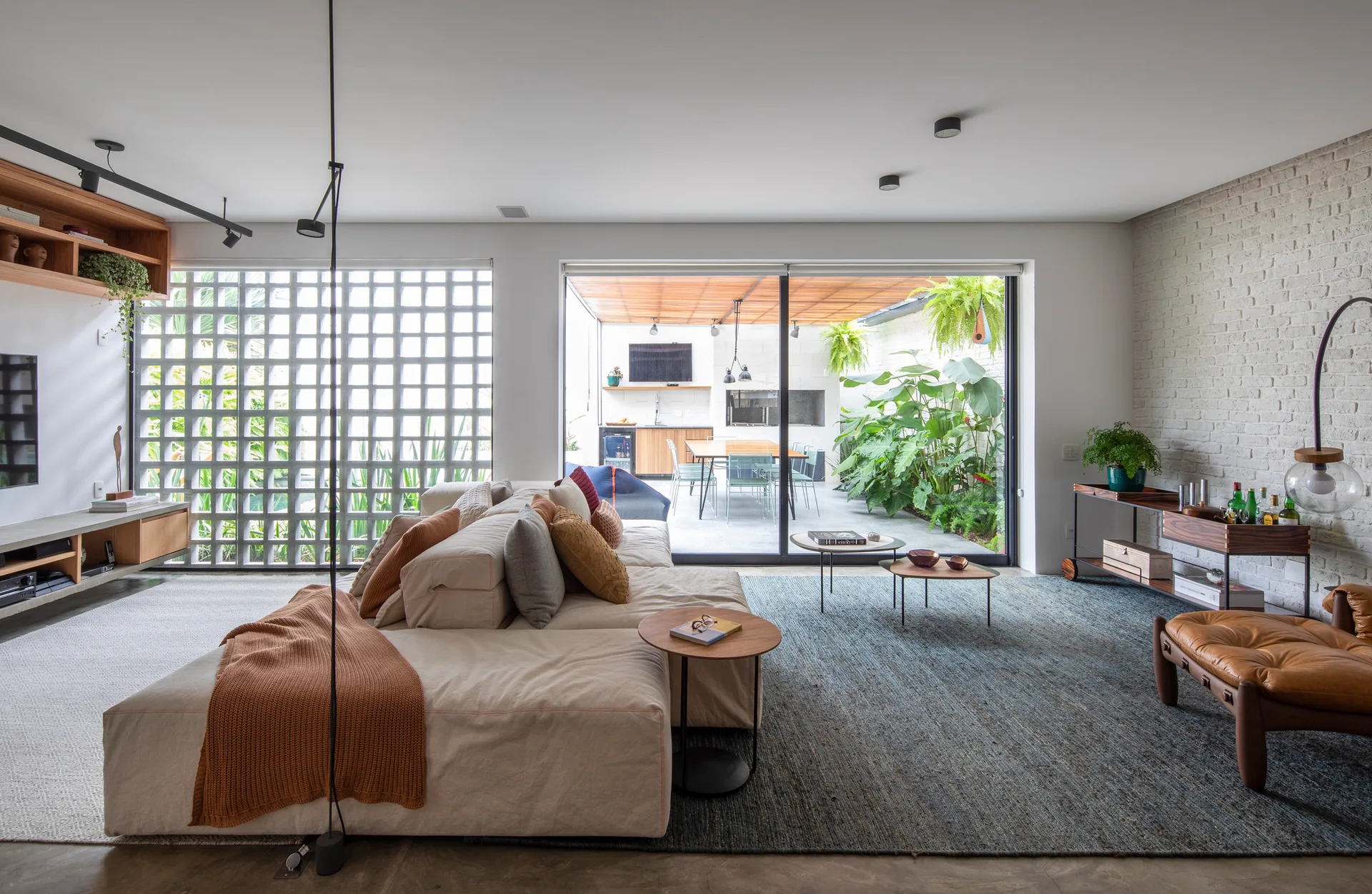 A modern open-plan living room features a cream sectional sofa, a textured blue rug, and a geometric cobogó screen next to a sliding glass door.