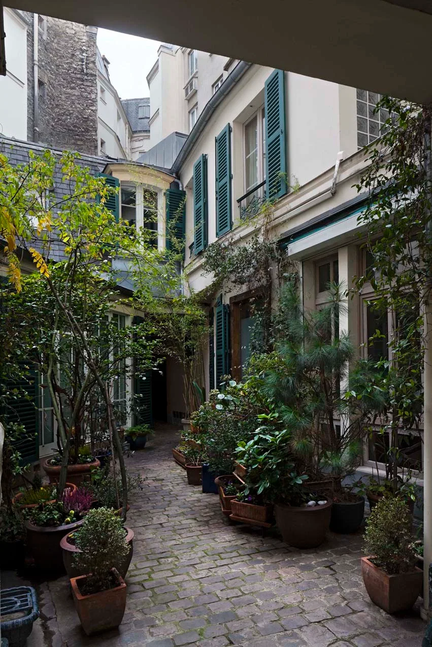 A stone courtyard with green window shutters, cobblestone paving, and terracotta planters filled with plants.