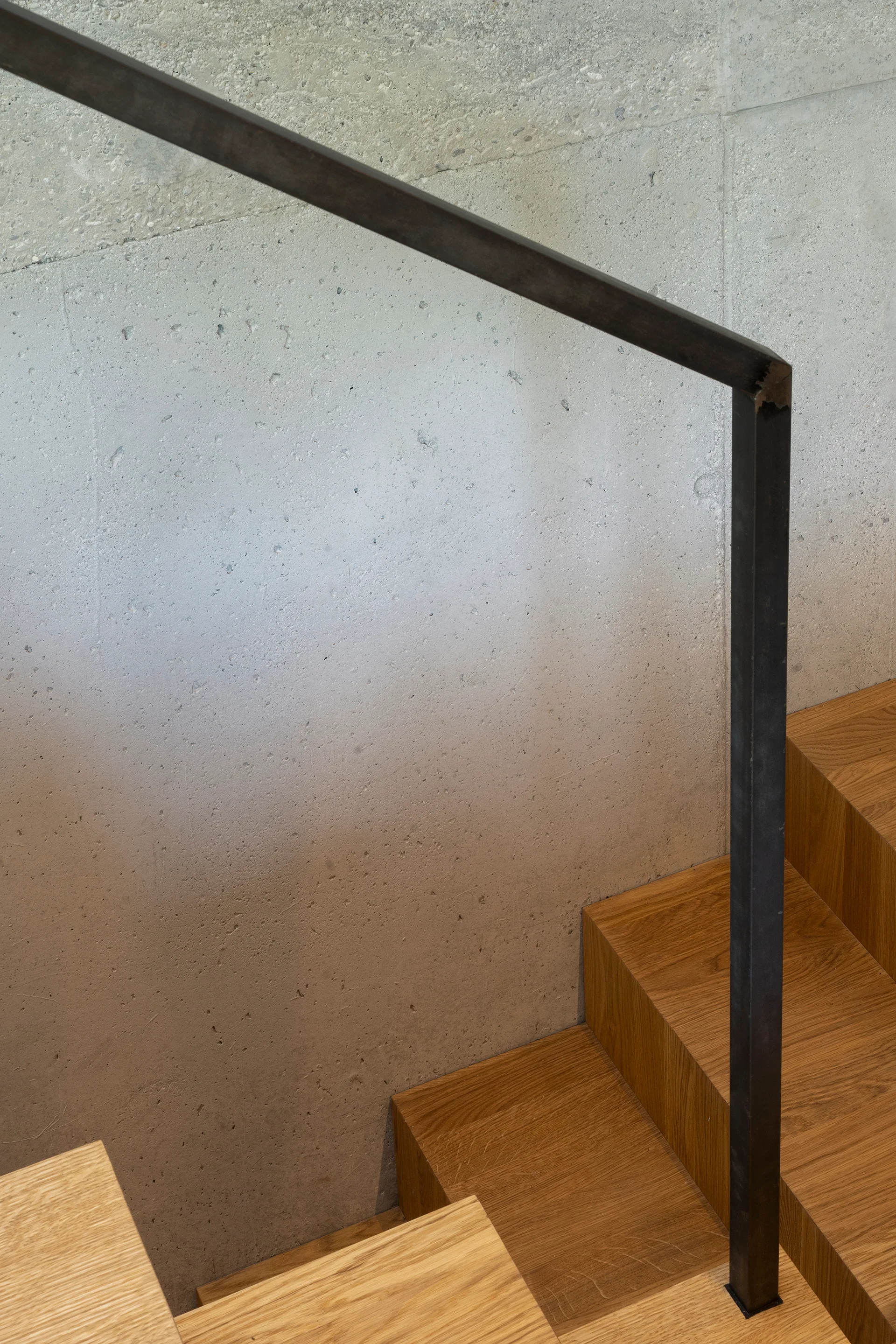 A modern staircase features warm wooden treads and a slim black metal handrail set against a textured light gray concrete wall.