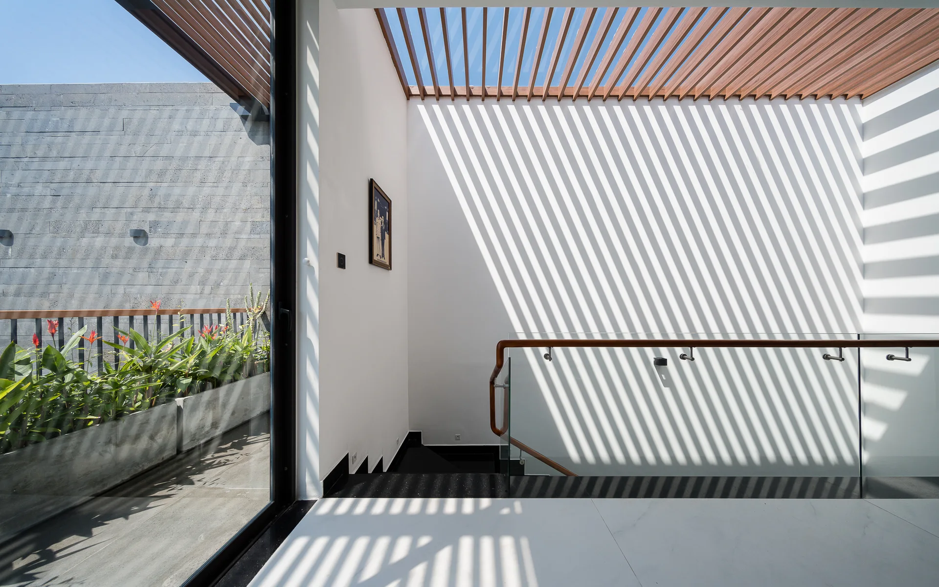 A bright staircase landing features a wooden slatted ceiling casting striped shadows on white walls next to a glass railing with a wood handrail.