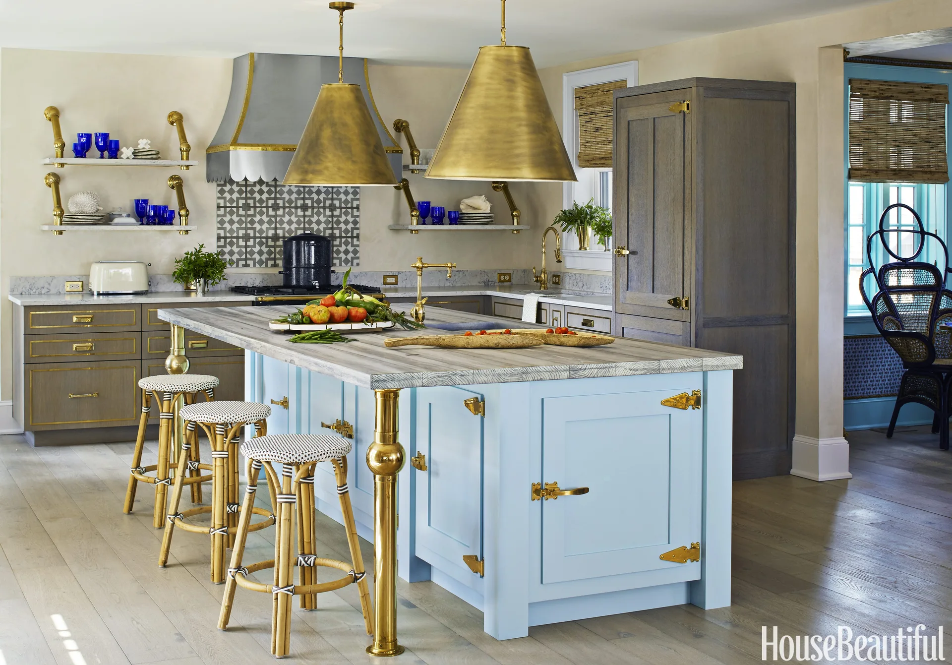 A kitchen features a light blue island with brass hardware, oversized brass pendant lights, bamboo barstools, and wood cabinetry.