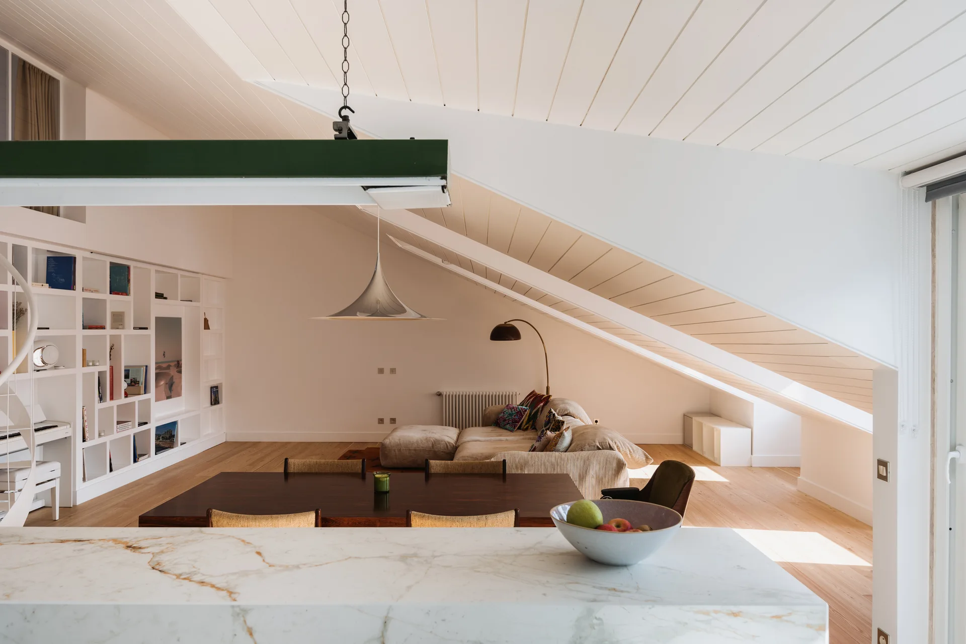 An open-plan attic living room featuring a slanted plank ceiling, a low beige sectional sofa, a dark wood dining table, and built-in white bookshelves.