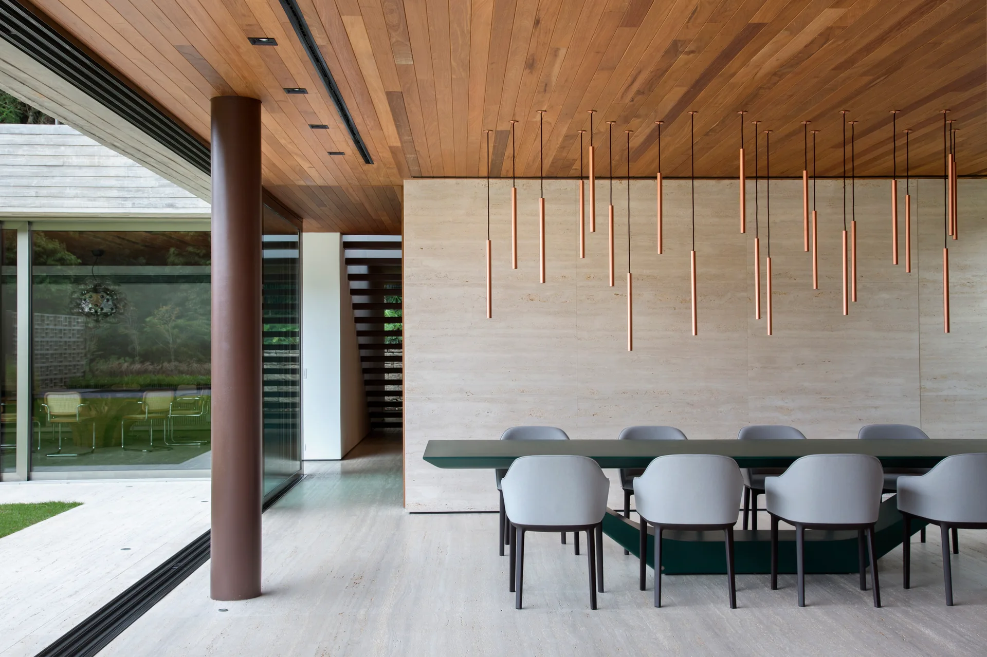 A minimalist dining room featuring a long green table, gray chairs, a travertine stone wall, and a cluster of slender copper pendant lights.