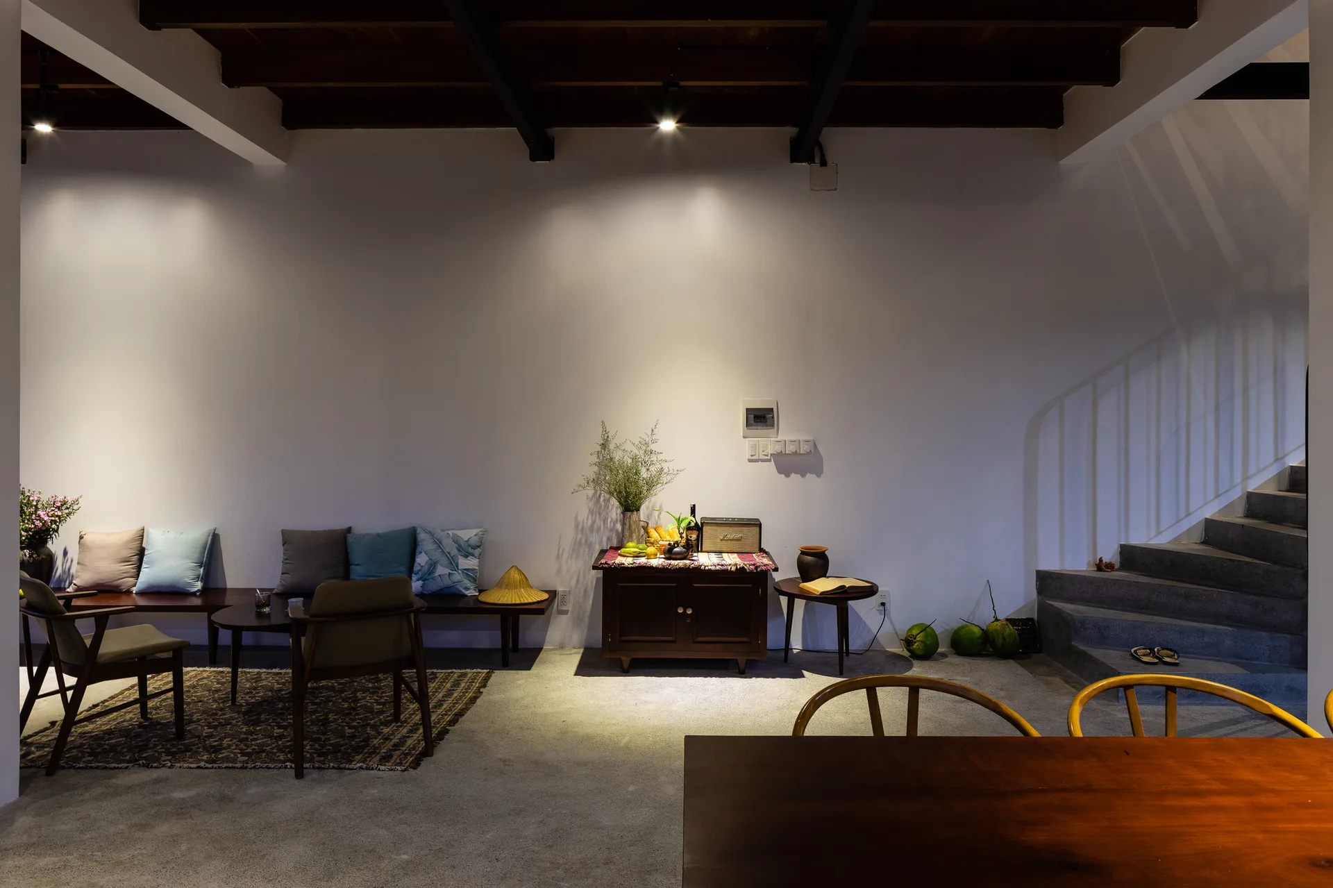 A minimalist living area features concrete floors, a concrete staircase, a dark wood bench sofa, and exposed ceiling beams under warm spotlights.
